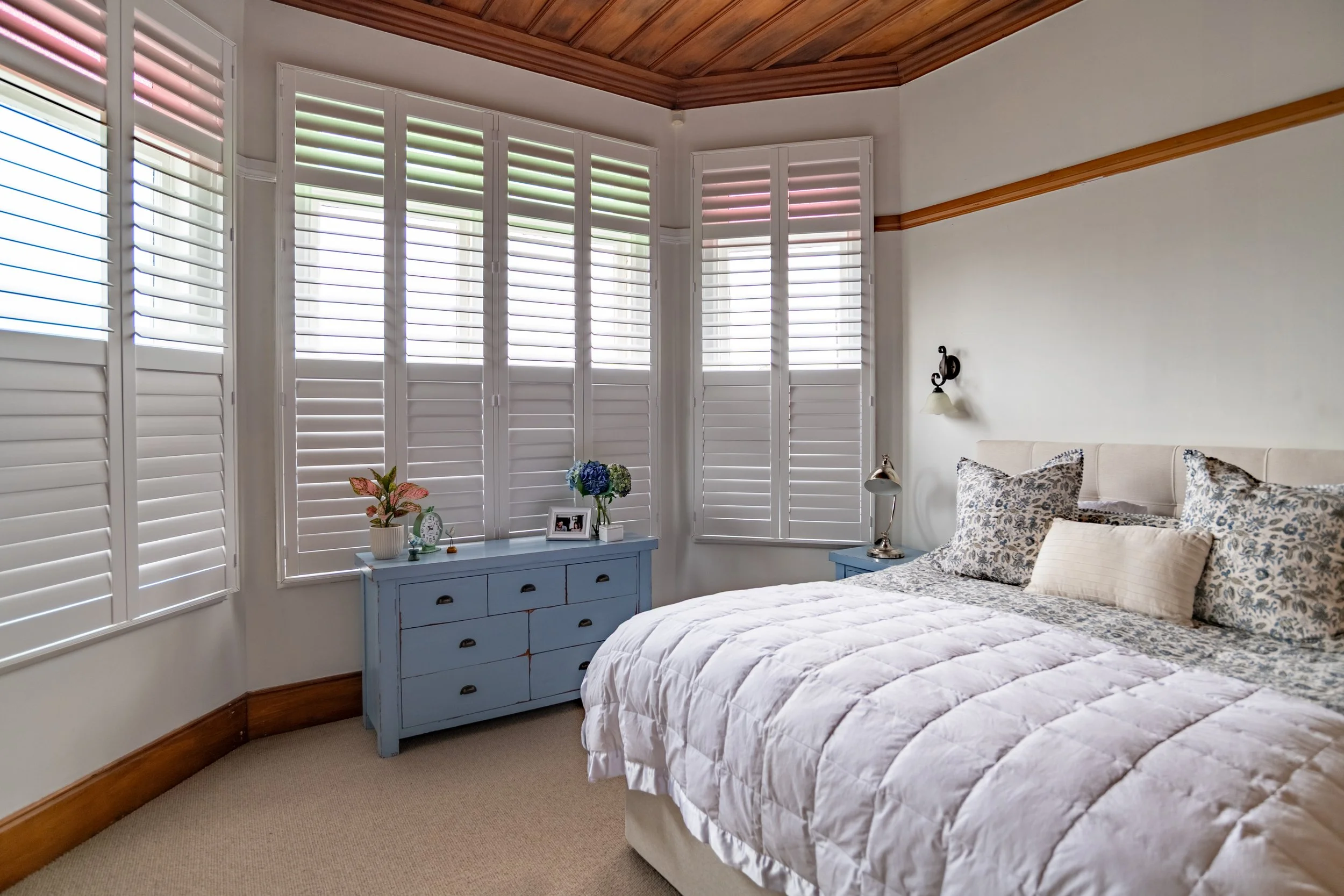 A cozy bedroom with a bed, patterned pillows, a nightstand with a lamp, and a blue dresser with plants and picture frames near window with white plantation shutters.
