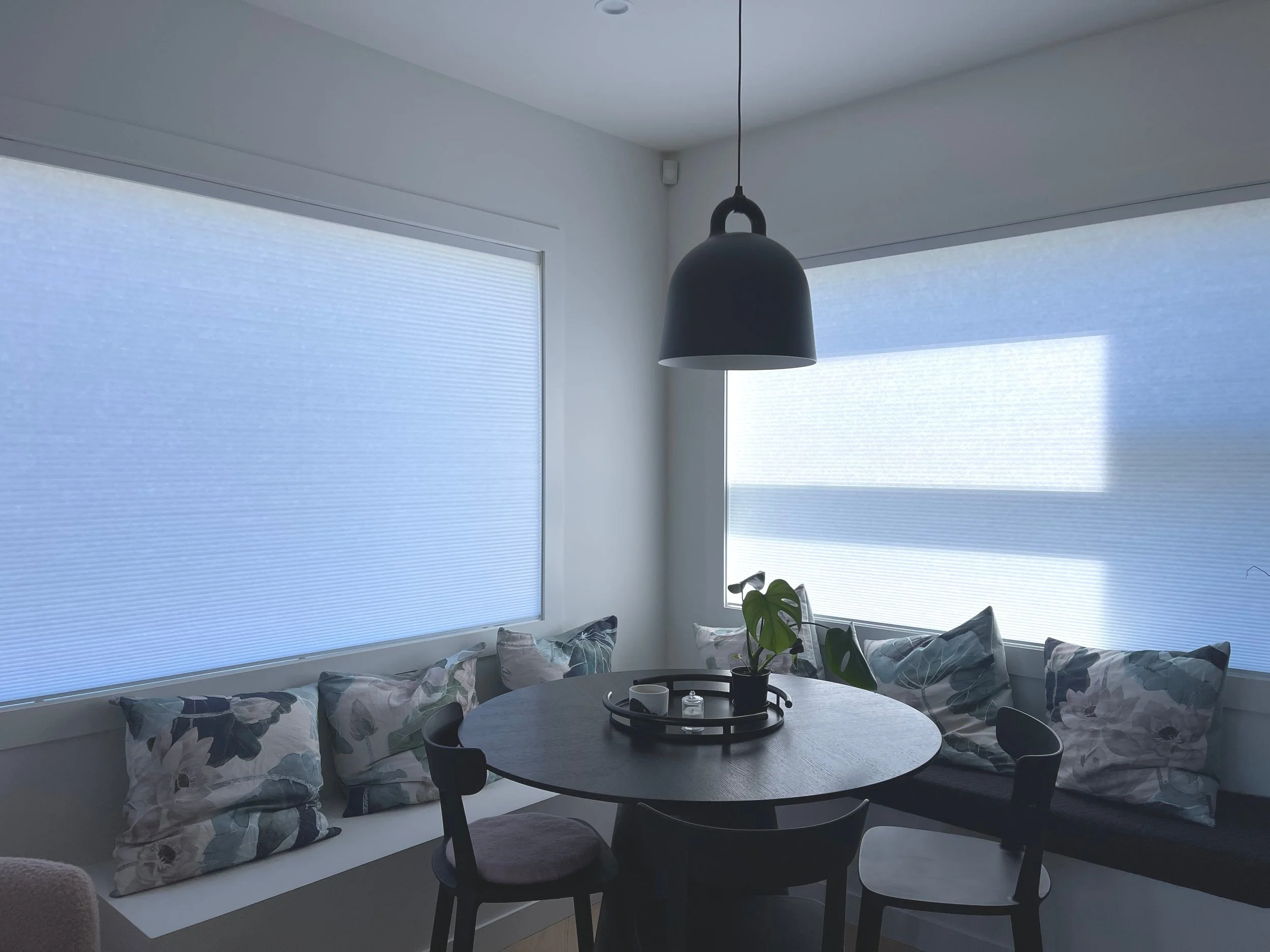A dining nook with a round black table, surrounded by black chairs with one cushion on each, a potted plant in the center, and windows with blue blinds letting in soft light.
