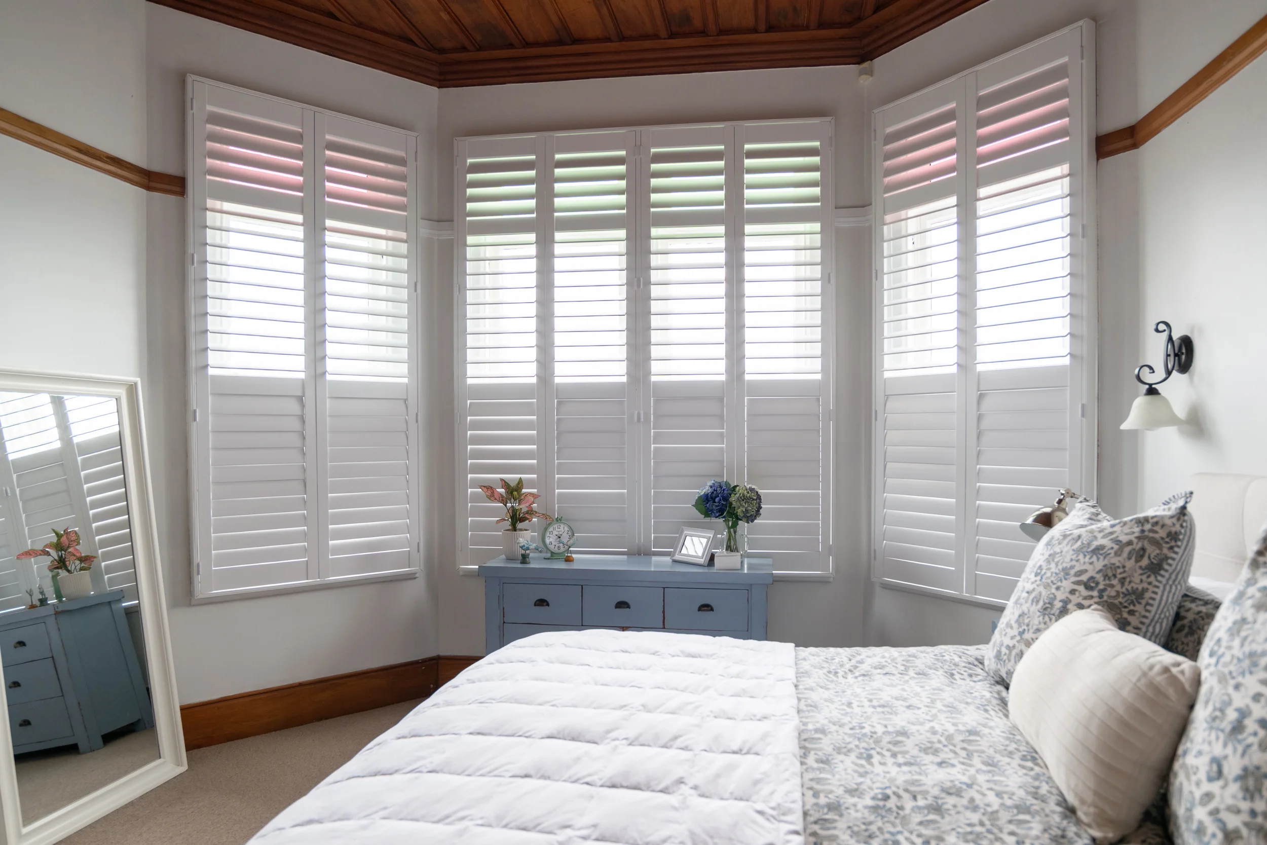 A bedroom with a bed, pillows, a blue dresser with flowers and framed photos, a standing mirror, white plantation shutters on multiple windows, a wall-mounted lamp, and wooden ceiling and trim.