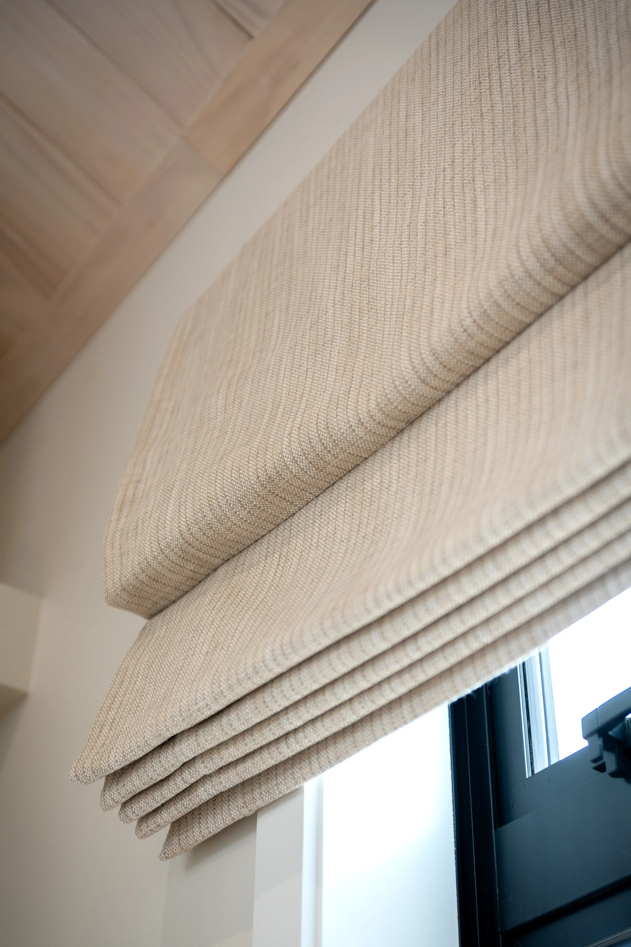 Close-up view of beige pleated window Roman shades covering a window, with a wooden ceiling and white wall nearby.