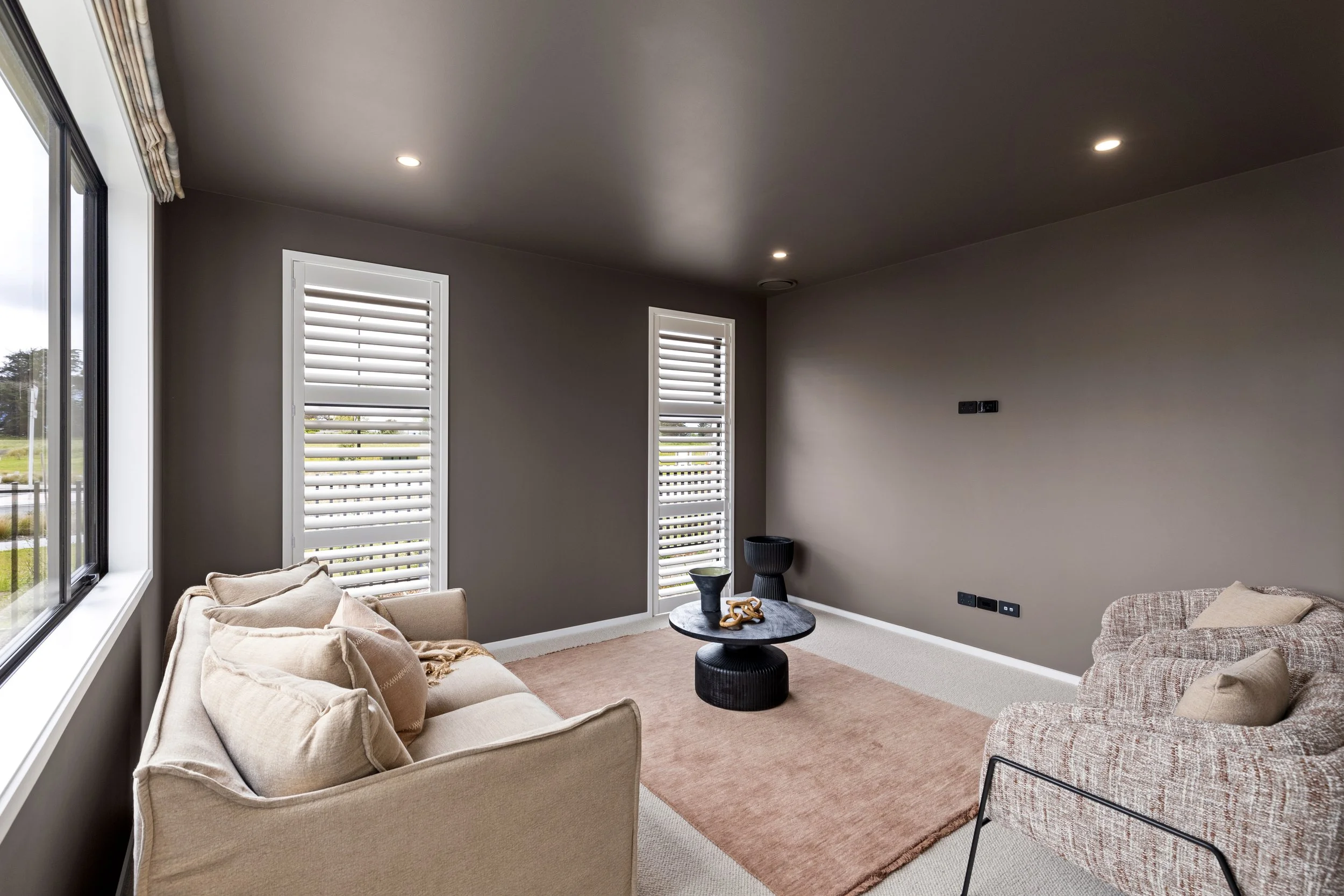 Modern living room with beige sofa, patterned armchair, black coffee table, blush area rug, gray walls, white window shutters, and large window with view outside