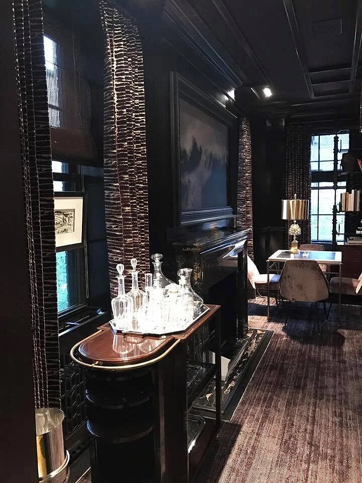 A cozy, elegant room with black walls, tall windows with patterned curtains, a fireplace, a bookshelf, a small round table with a gold lamp, and a bar cart with glass decanters.