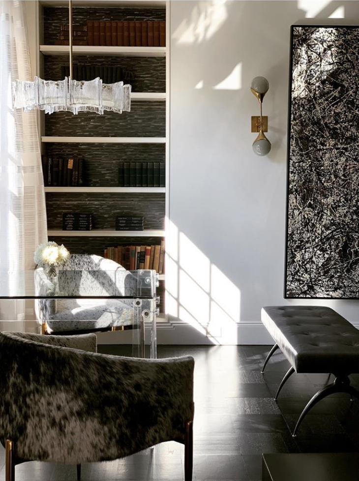 Bright, modern living room with sunlight coming through large window, featuring a glass table, plush armchairs, a black and white artwork, and built-in bookshelves with books.
