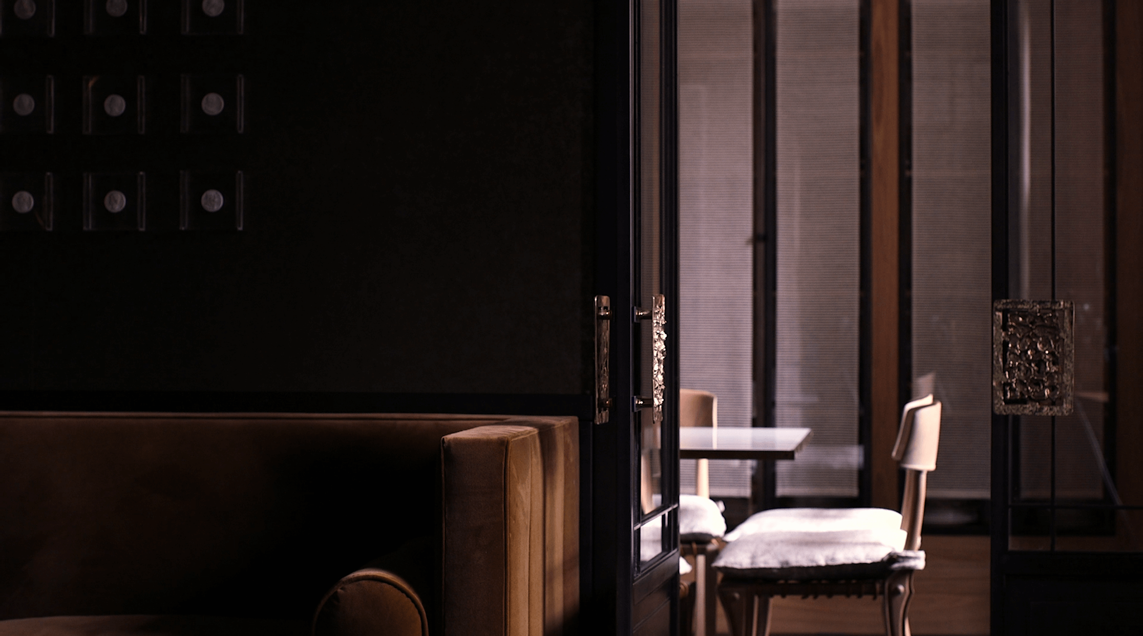 A dimly lit interior of a restaurant or cafe with a private dining area separated by black-framed glass doors. Inside, there are white chairs and a table, with textured, dark-colored wall panels.