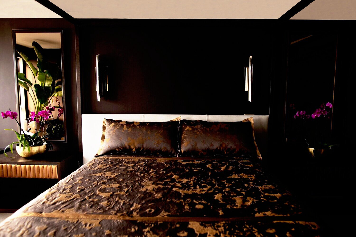 A bedroom with dark walls and a bed with brown and gold bedding, flanked by side tables with potted purple orchids, and a large mirror and window with blinds on the left side.