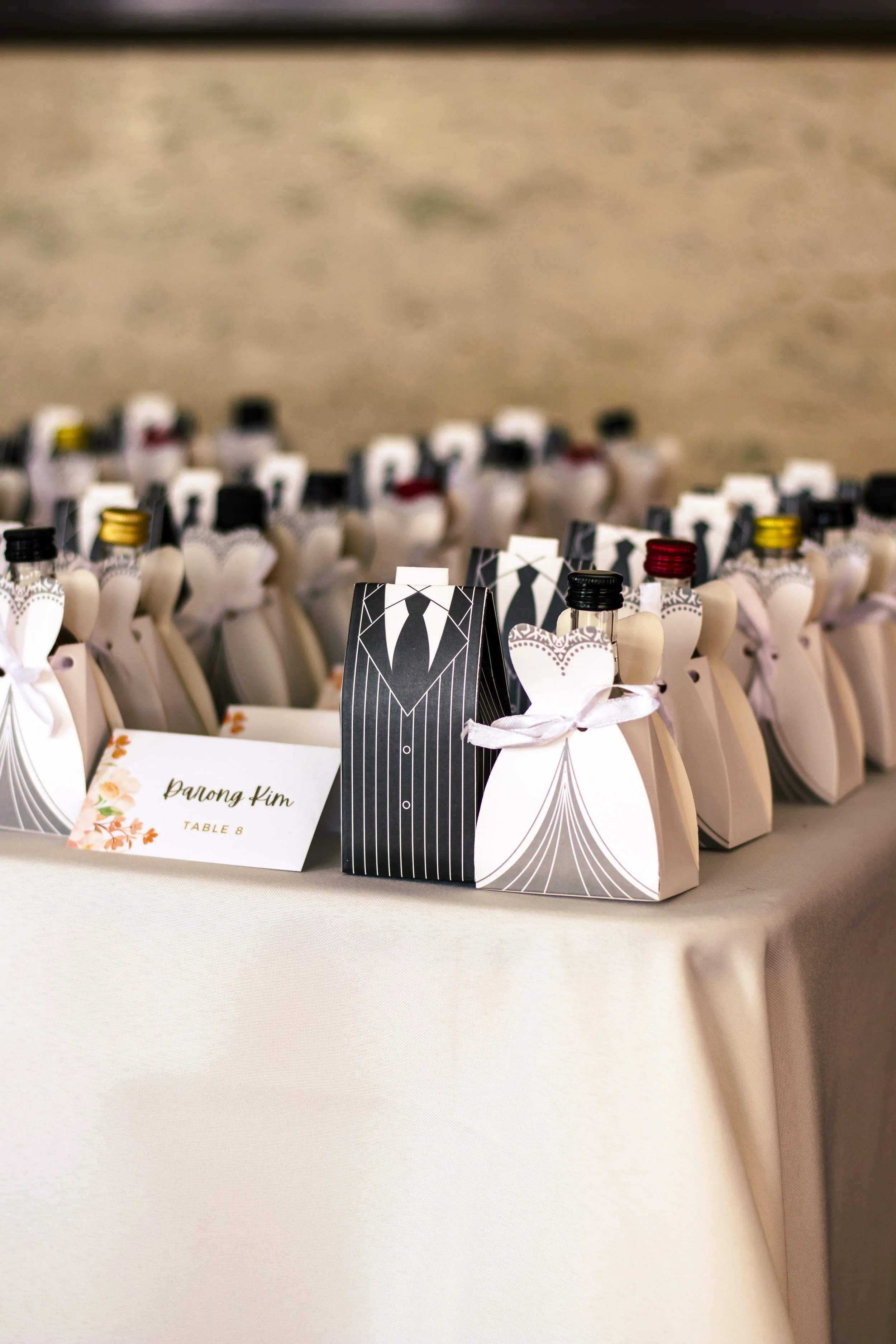 Miniature wine bottles in tuxedo and dress-themed packaging, arranged on a table with a name card for guest seating.