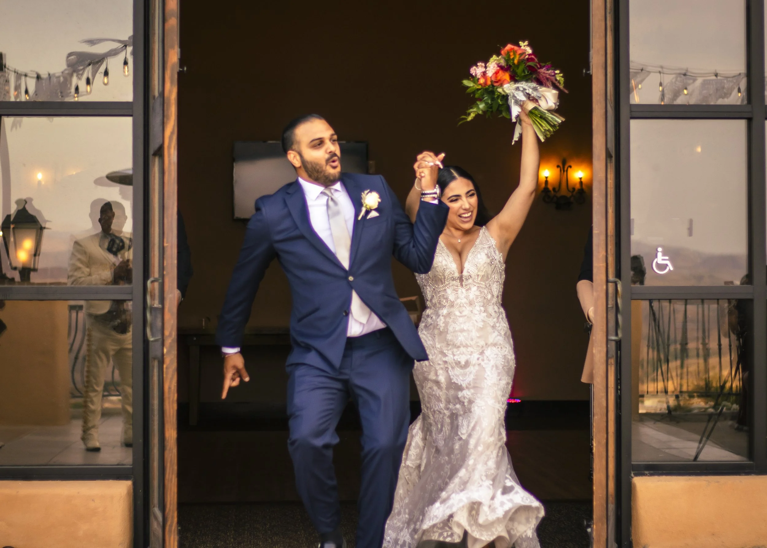 A joyful couple in wedding attire exiting a building, with the bride holding a bouquet and both raising their hands in celebration.