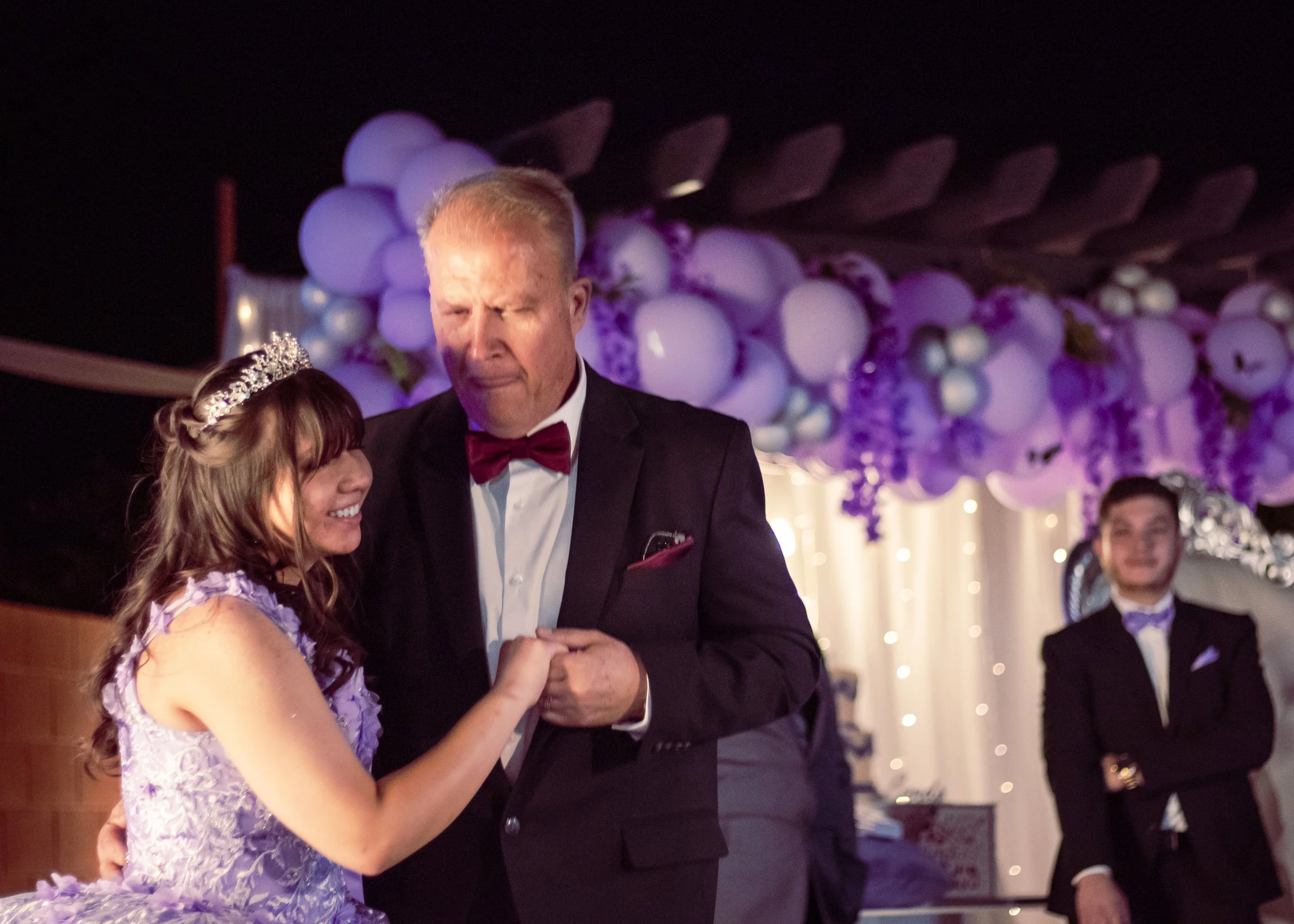 A father and daughter dancing at a wedding reception, with the daughter wearing a lavender dress and tiara, and the father wearing a tuxedo with a red bow tie, pink pocket square, and a white shirt. The background features purple balloon and floral d