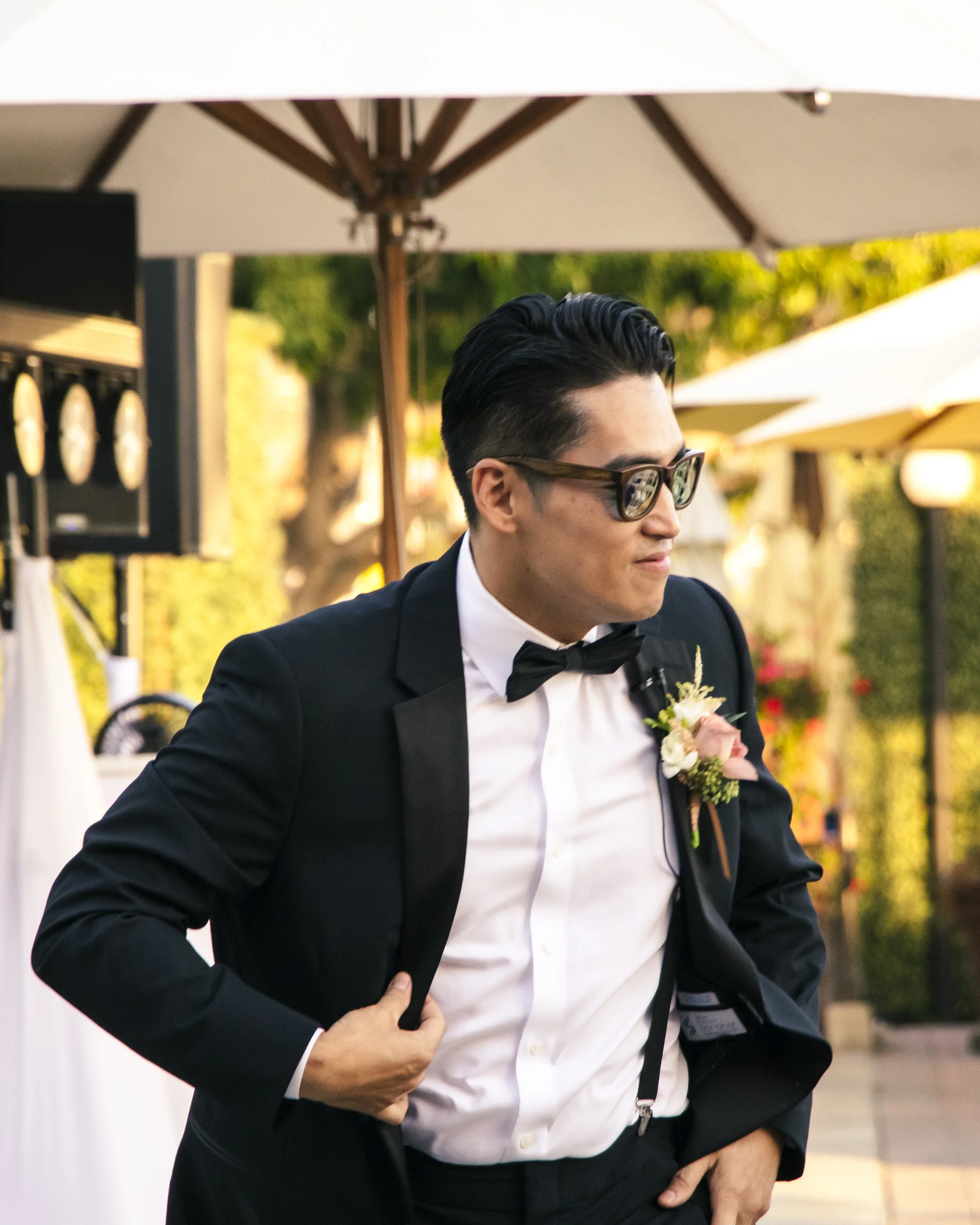 Man in formal attire with sunglasses, adjusting suit jacket outdoors under umbrellas.