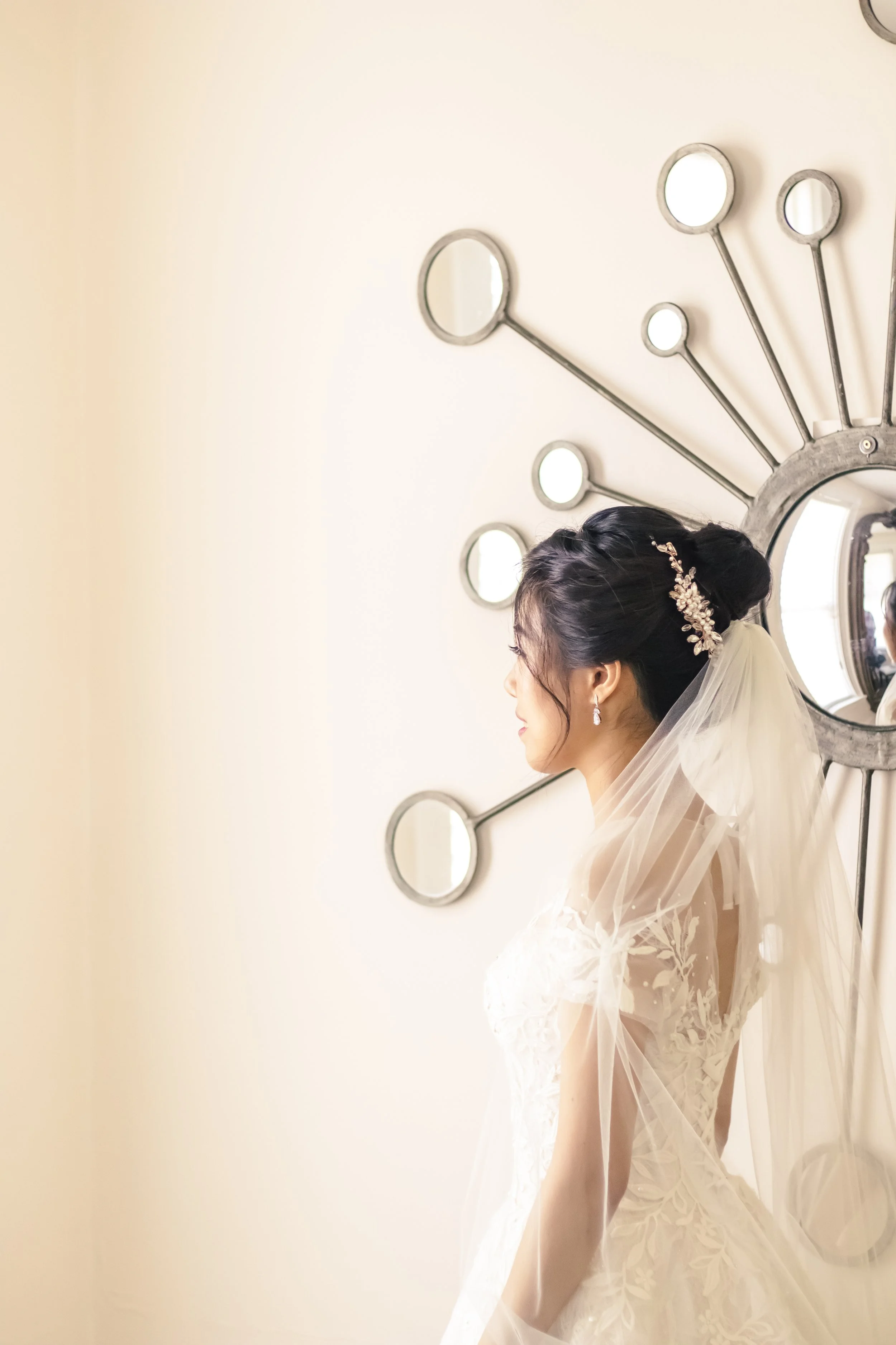Bride in white lace gown with long veil standing sideways, gazing towards a decorative wall mirror with multiple circular elements.
