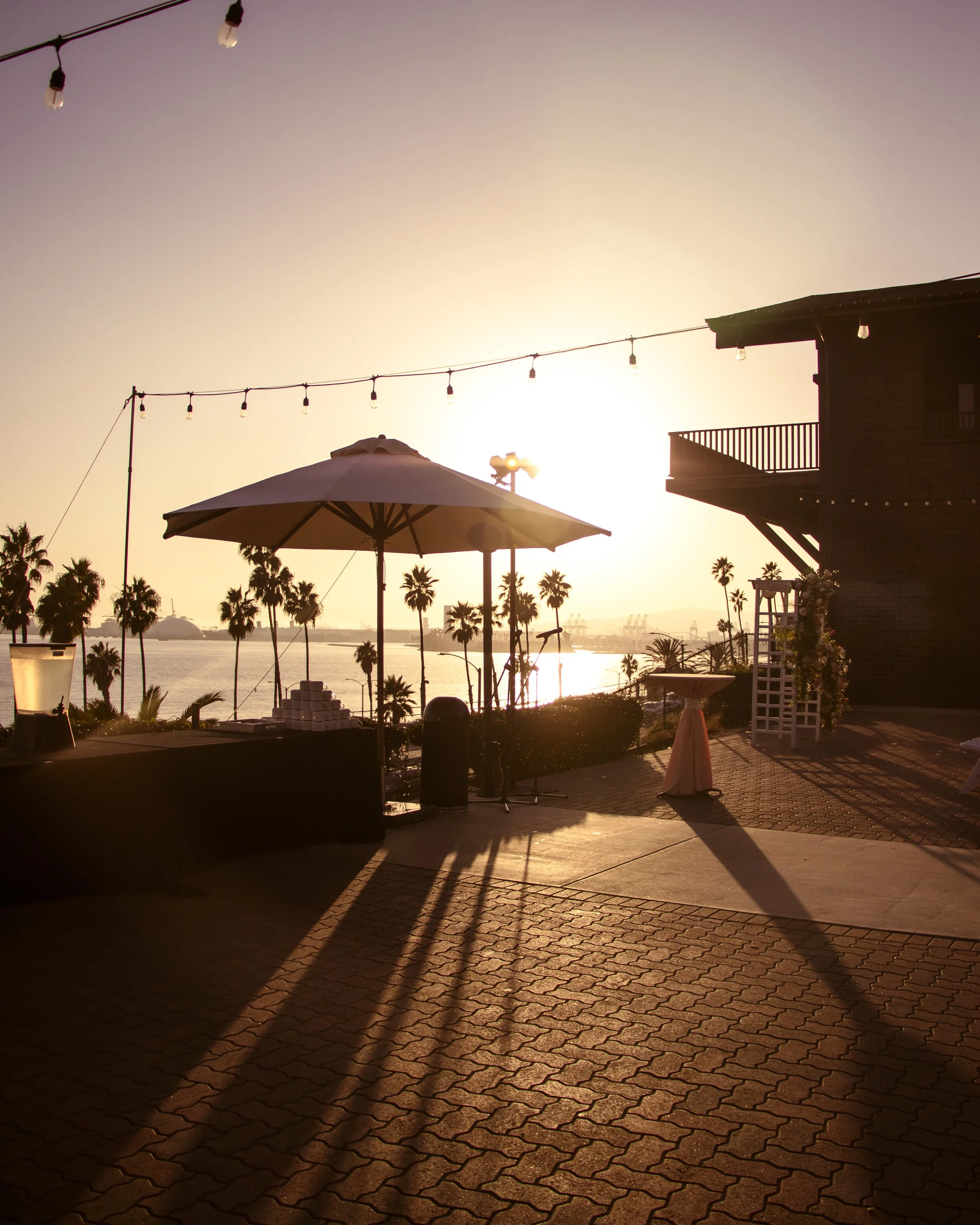 Outdoor patio at sunset with string lights, palm trees, and an umbrella. Silhouetted building and ocean view in the background.