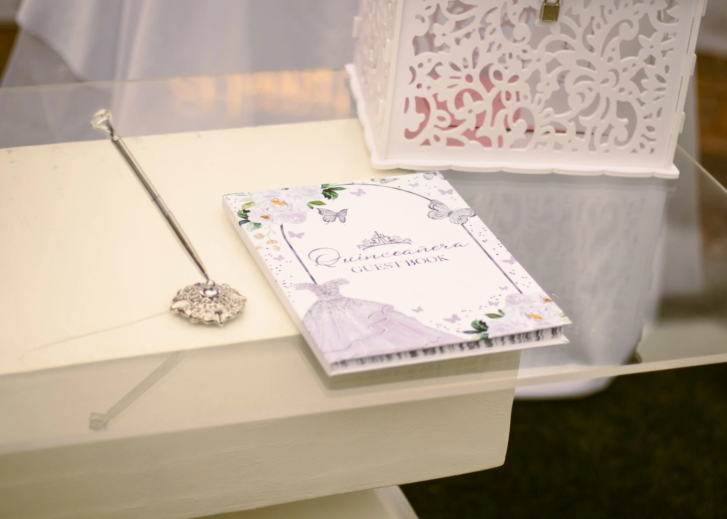 Guest book labeled 'Queenara Guest Book' with floral and butterfly design, a silver pen with decorative holder, on a white table near a white decorative box.