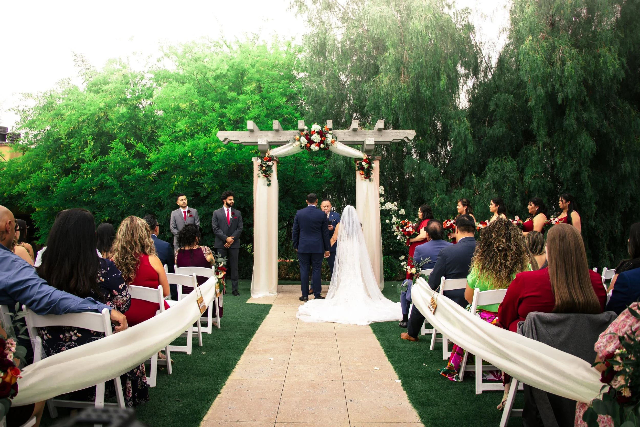 Outdoor wedding ceremony with bride, groom, officiant, bridesmaids, groomsmen, guests, floral arch, and greenery backdrop.