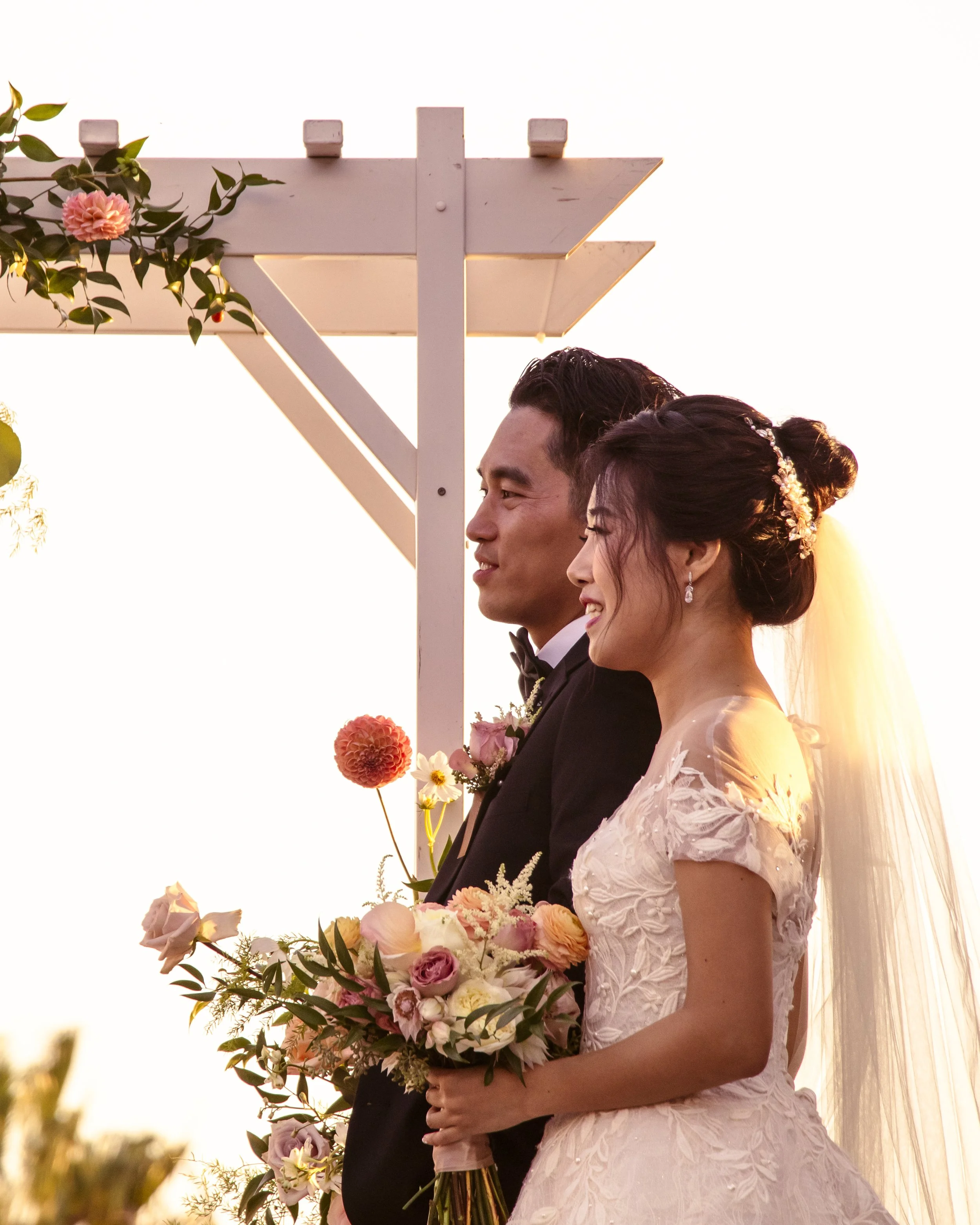 A bride and groom stand together under a floral-decorated pergola, the bride holding a bouquet of flowers. The sunlit scene suggests a wedding ceremony.