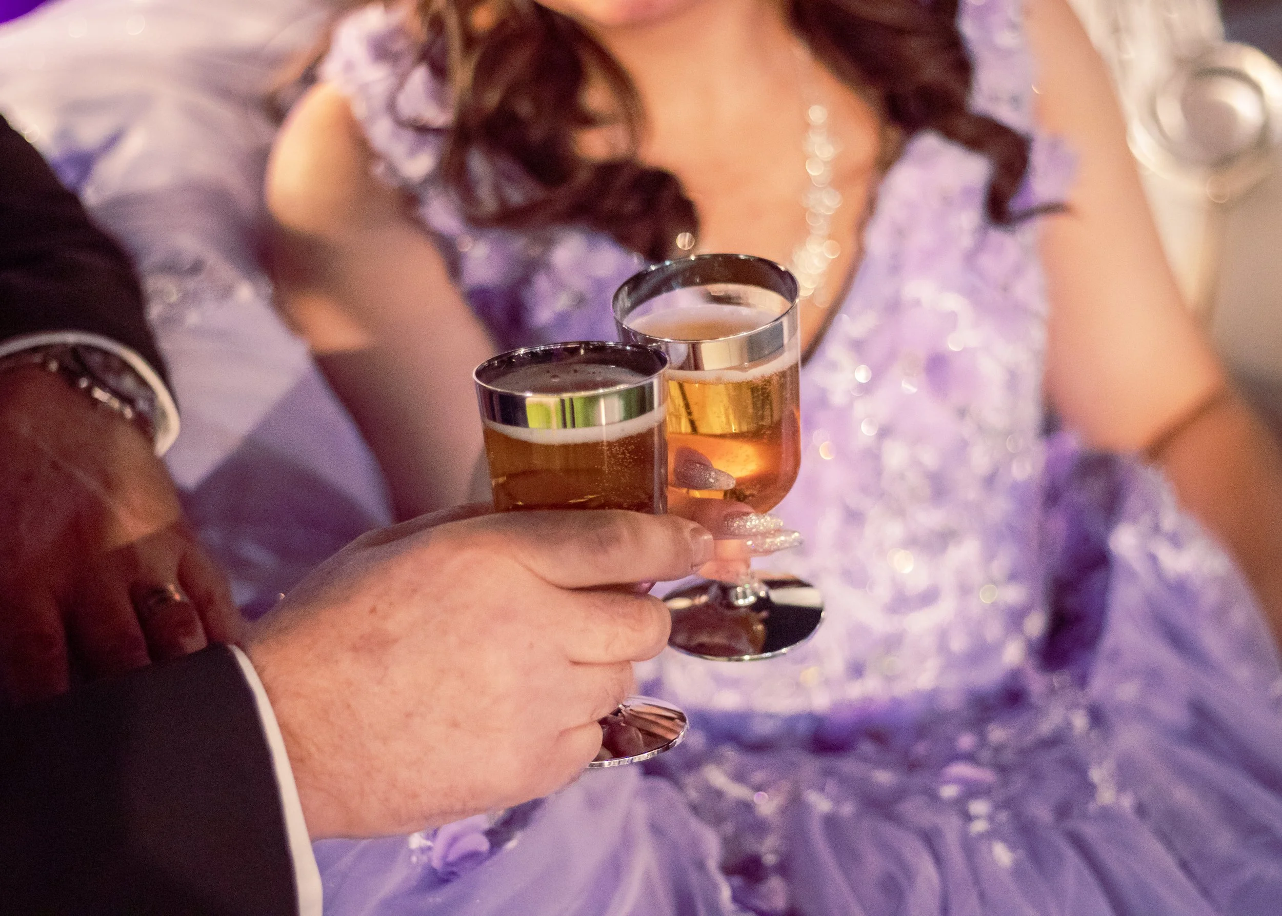 Close-up of a man and woman holding champagne glasses in a toast. The woman is wearing a fancy purple dress and jewelry, the man is in a dark suit. The background suggests a celebration or special occasion.