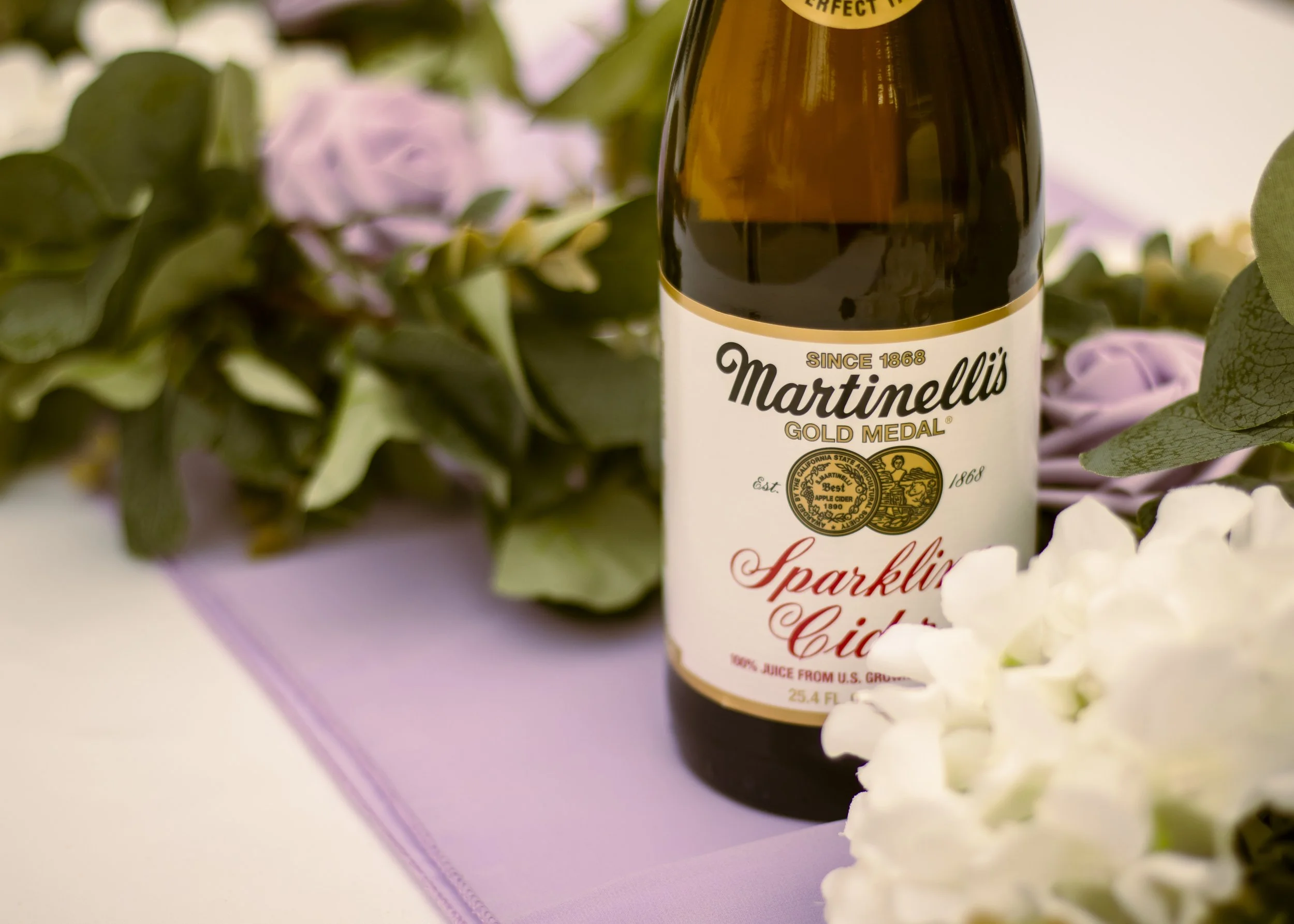 A bottle of Martinelli's Sparkling Cider placed on a purple cloth with purple and white flowers and green leaves around it.