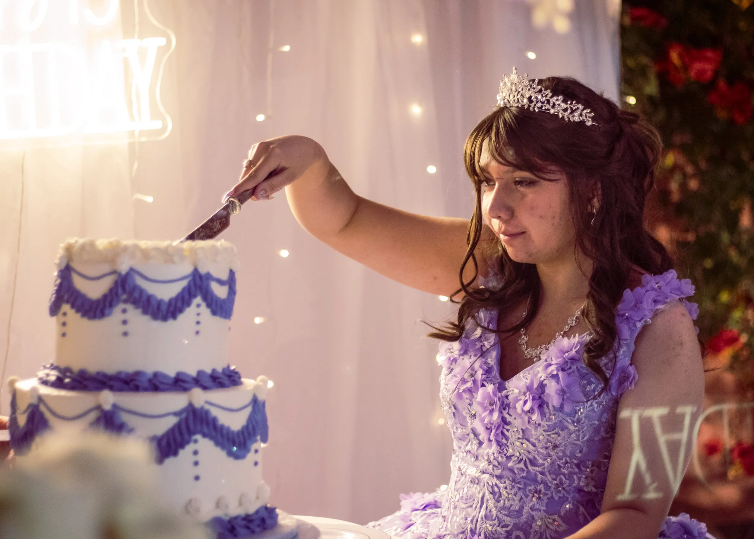 Young woman in a lavender dress and tiara cutting a decorated wedding cake.