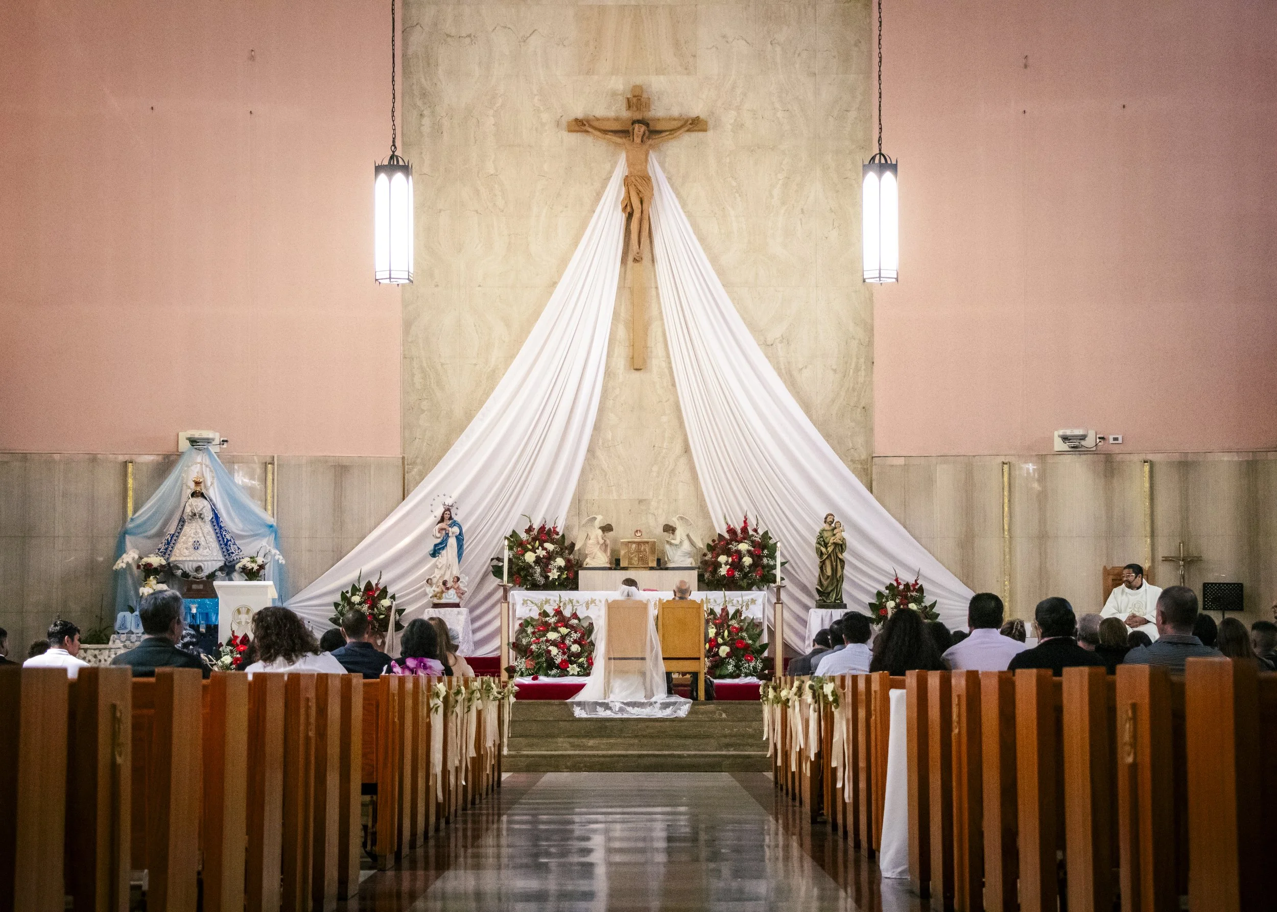 Interior of a church during a wedding ceremony, featuring pews, a couple at the altar, religious statues, and floral arrangements.