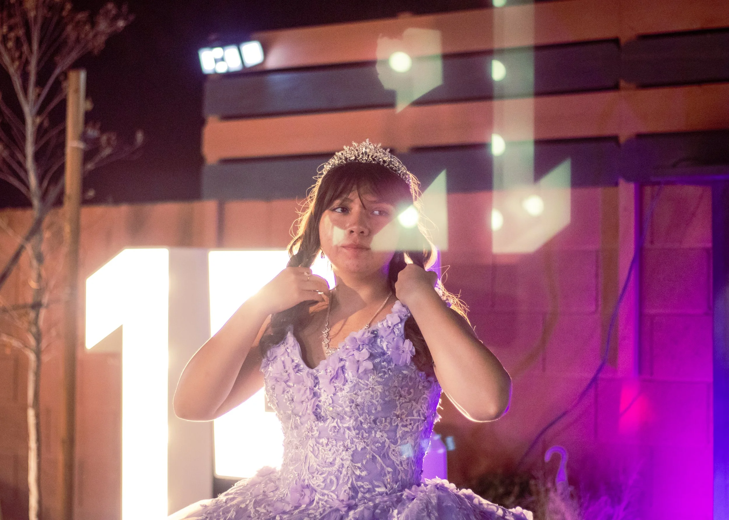 A young woman wearing a lavender lace dress and a tiara standing in front of colorful lights at an indoor event.