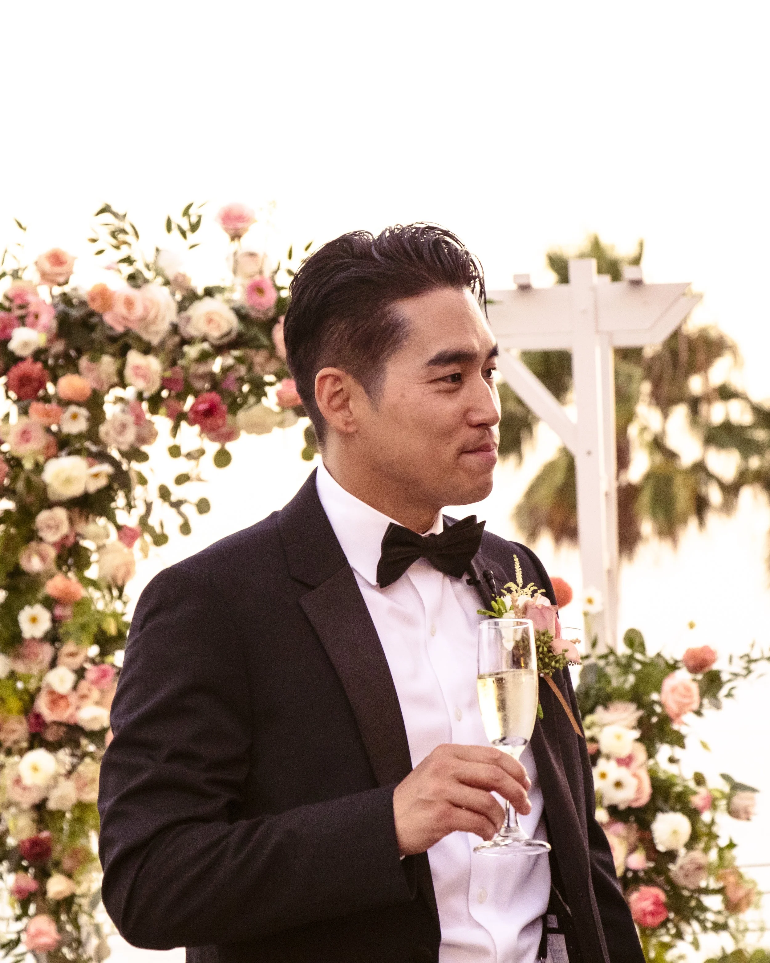 Man in a suit with a bow tie holding a champagne glass, standing in front of a floral backdrop.