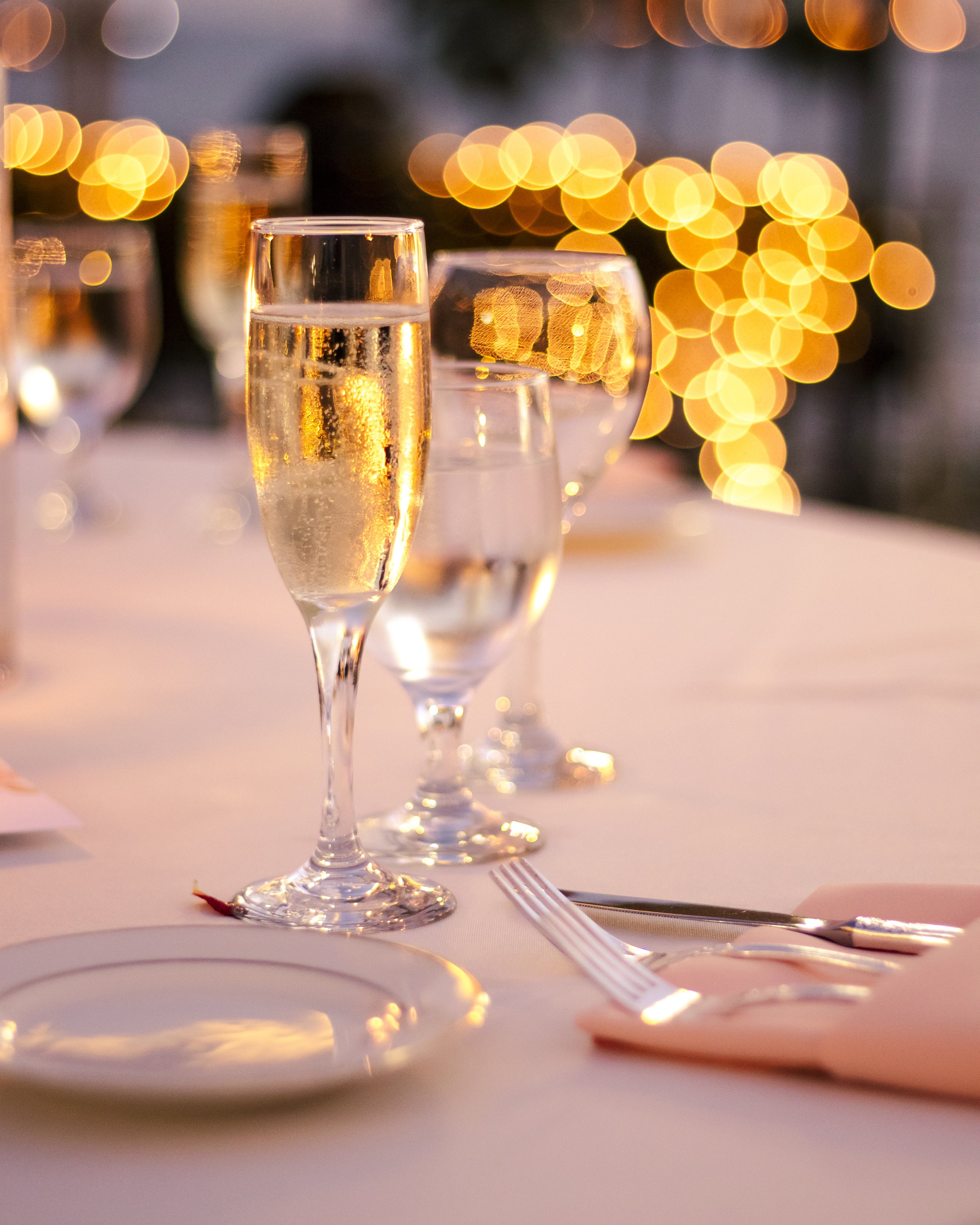 Close-up of a table setting with a champagne glass, water glasses, plate, and cutlery on a white tablecloth, with blurred golden bokeh lights in the background.