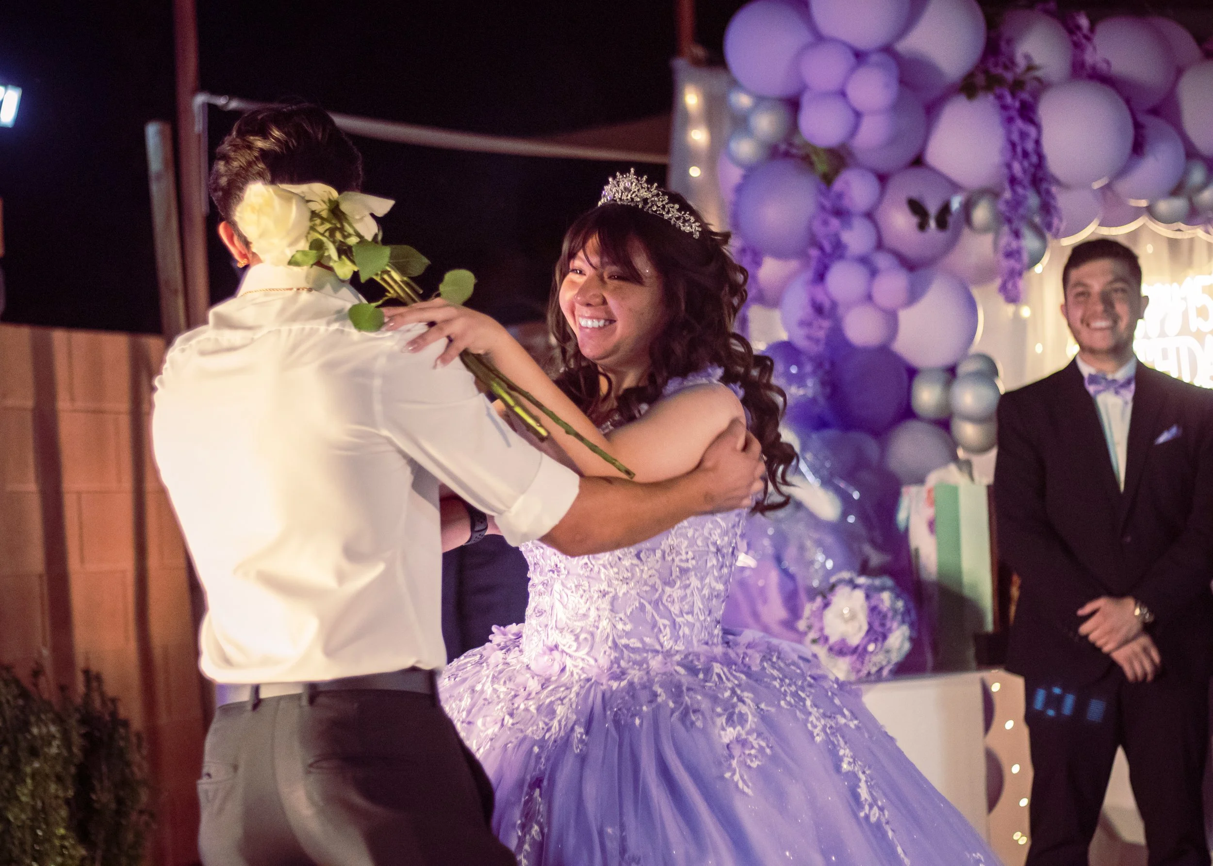 A woman in a purple wedding dress with a tiara is smiling as a man in a white shirt gives her a bouquet of white roses during a wedding celebration. A man in a black suit stands in the background near purple balloon decorations.