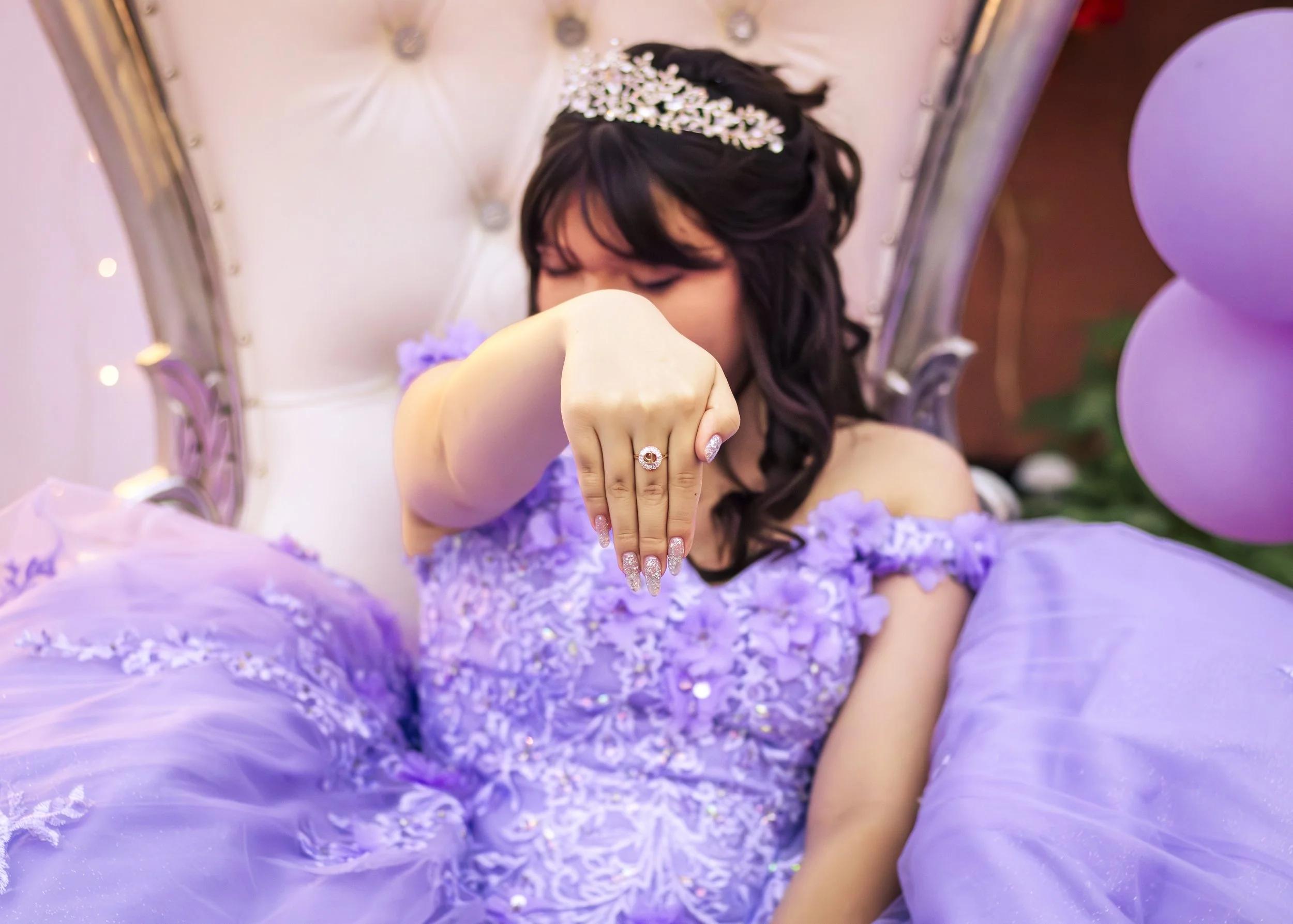 A young woman in a purple dress with floral lace details is sitting on a white ornate chair. She is wearing a tiara and displaying a ring on her finger, with her hand extended towards the camera. Pink balloons are visible in the background.