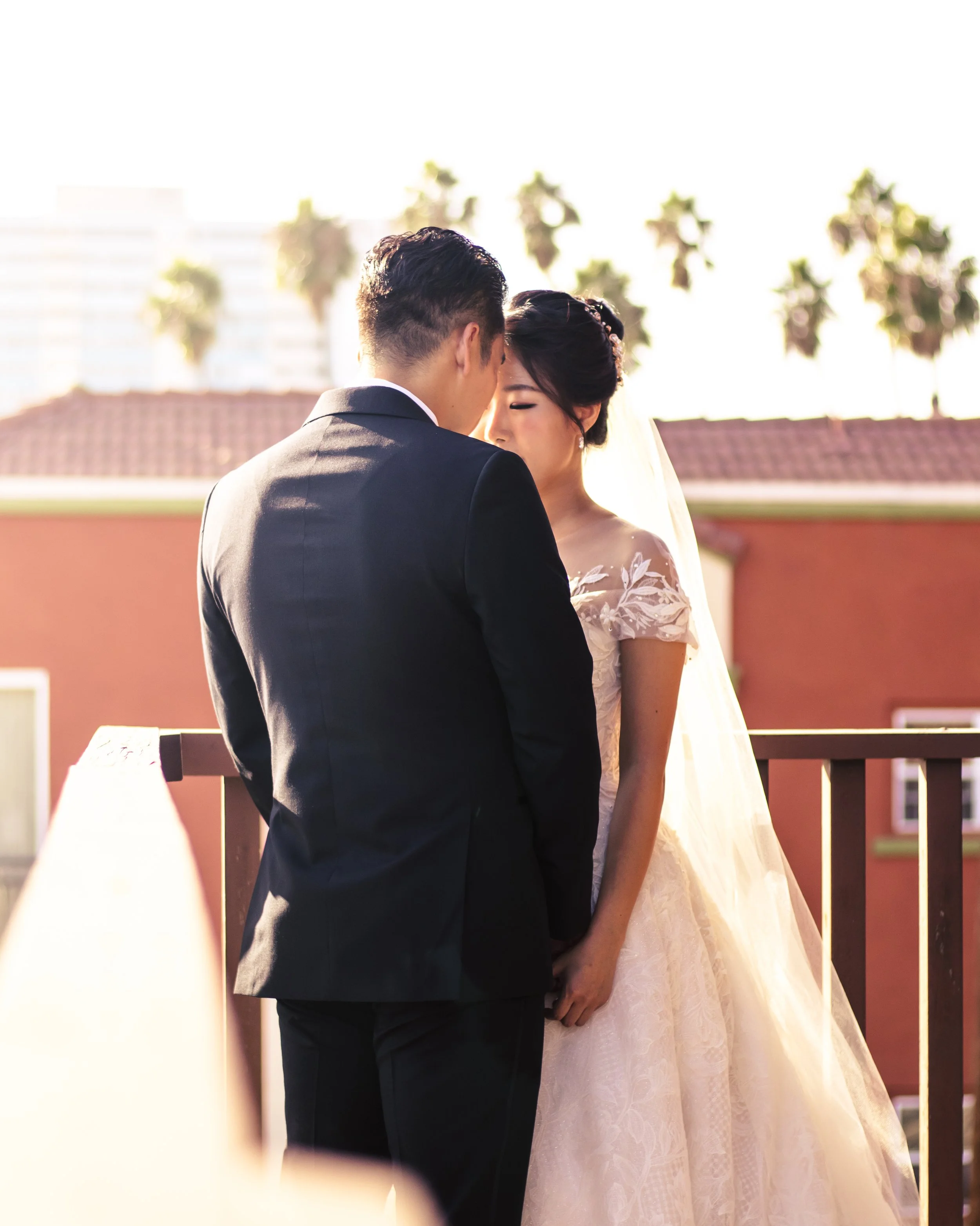 Bride and groom standing closely on a balcony with palm trees in the background.