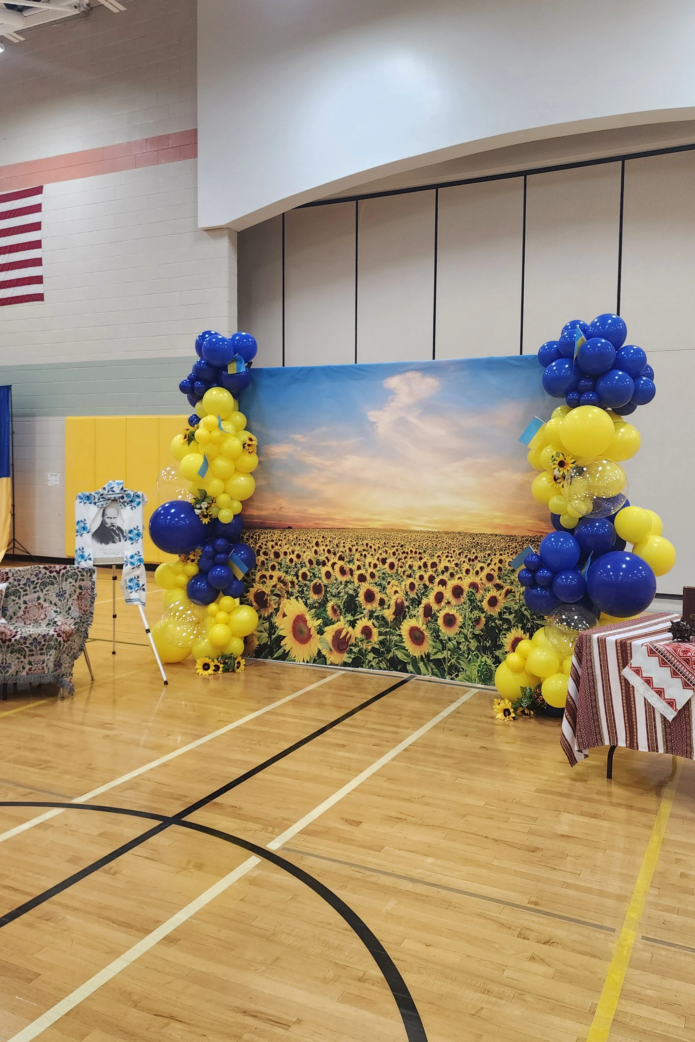 A sunflower field backdrop with yellow and blue balloon decorations on either side, set up in a gymnasium with wooden flooring.