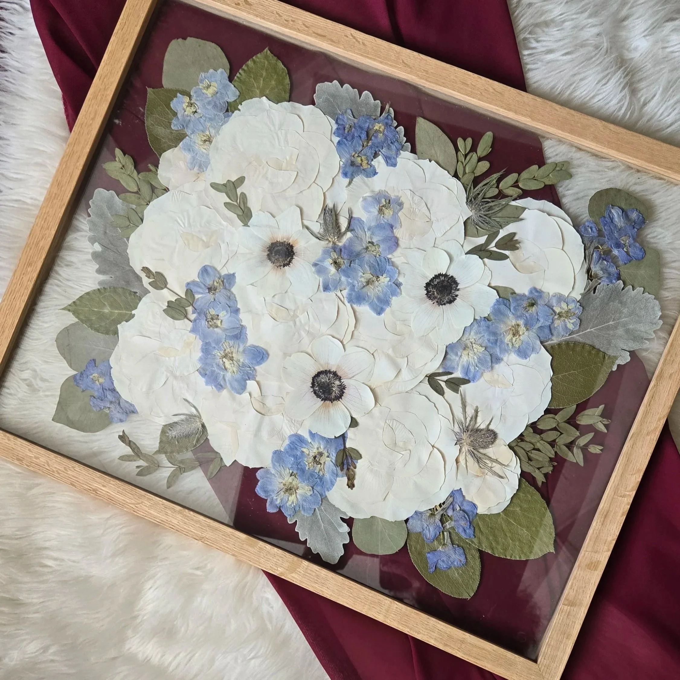 A pressed flower art piece featuring pink, white, and blue flowers with green leaves, framed in wood and resting on a pink fabric and white furry surface.