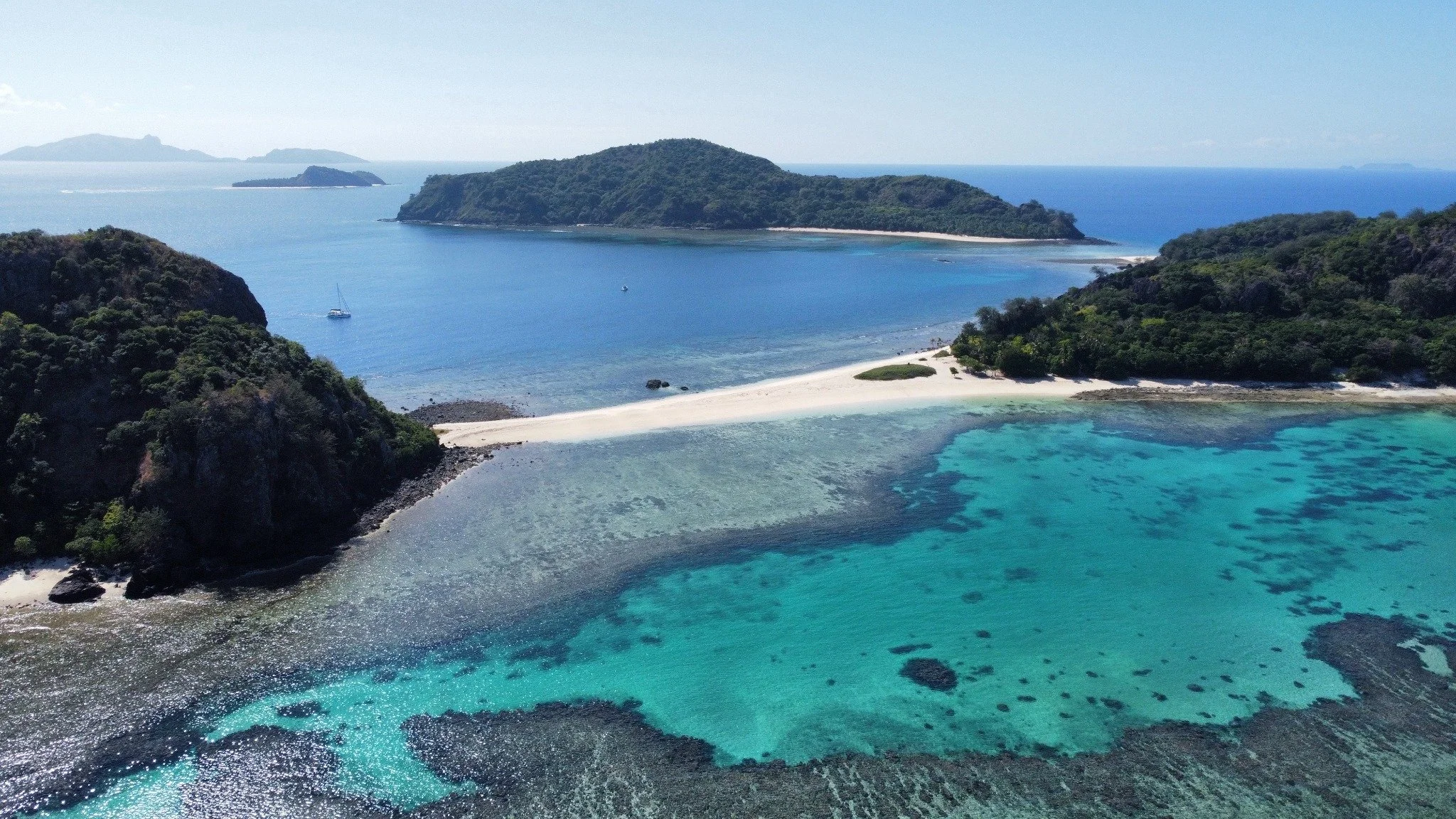 Aerial view of tropical islands, a sandy beach and an anchored yacht in turquoise reef waters.