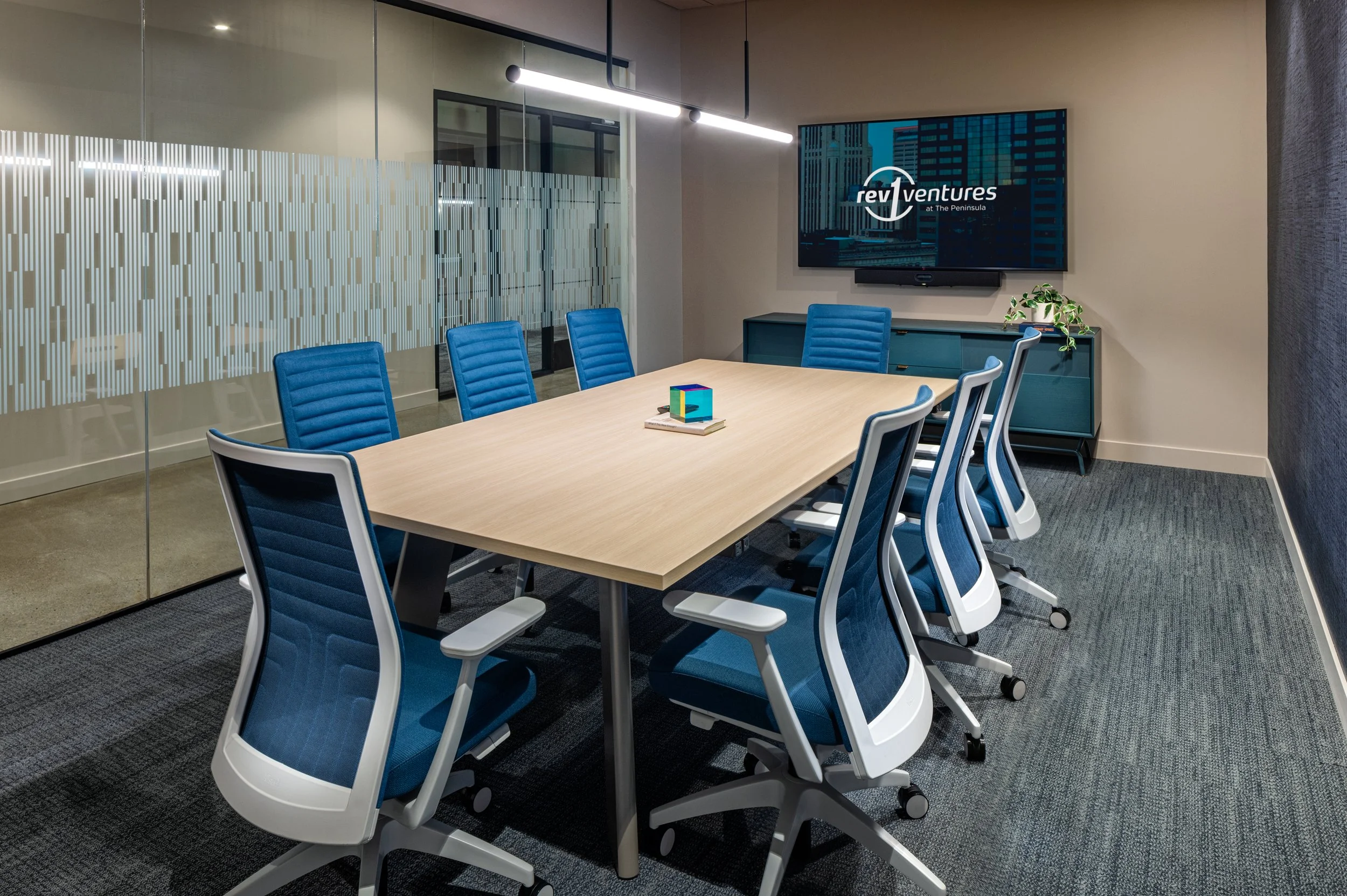 Modern conference room with a wooden table, ten blue and white office chairs, a TV screen displaying 'rev ventures,' a small cabinet with a plant, and a glass wall with frosted patterns.