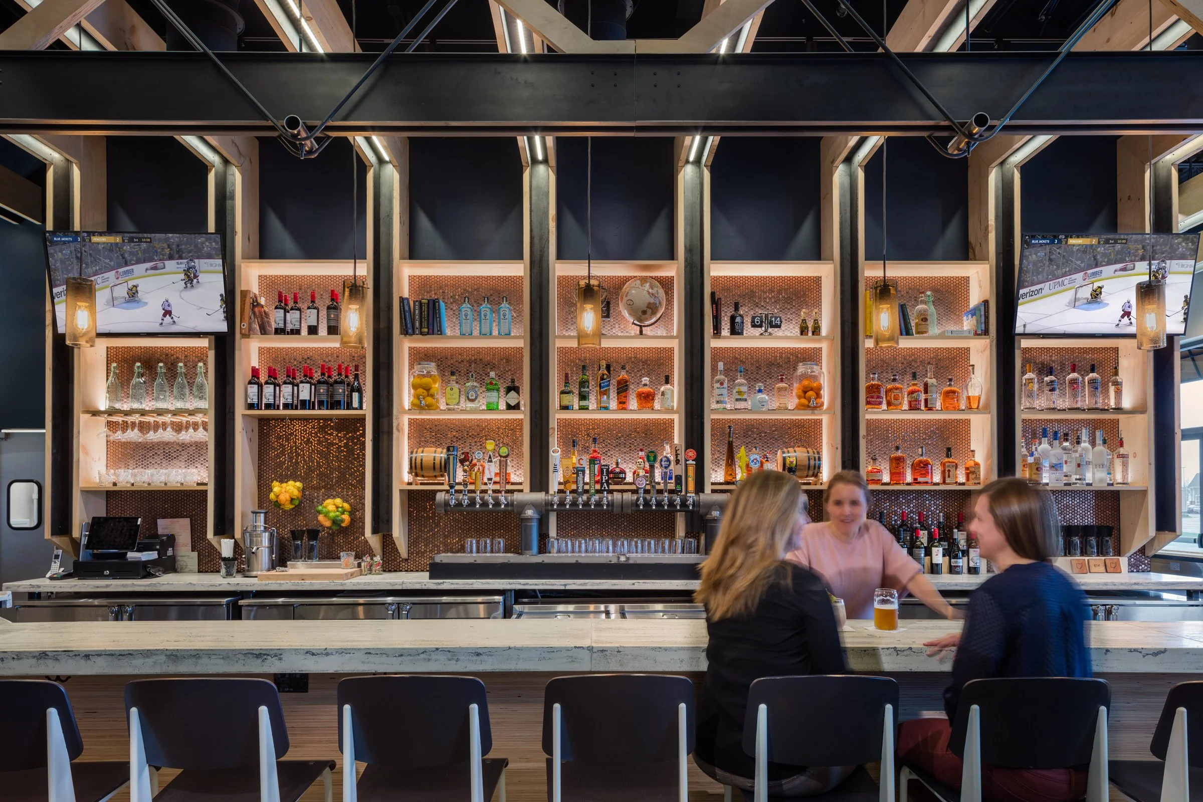 Interior of a modern bar with three women sitting at the countertop, bar shelves stocked with liquor bottles, two TV screens showing hockey games, and warm lighting above the bar.