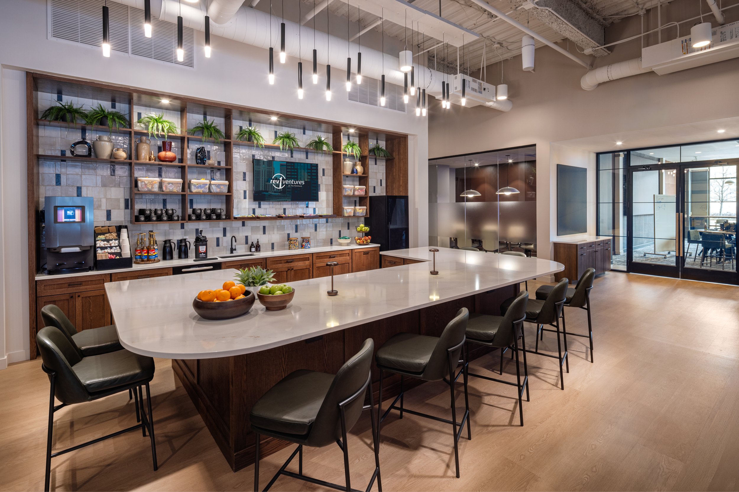 Modern break room with a large oval island table, chairs, a coffee station, and decorative plants on a back wall
