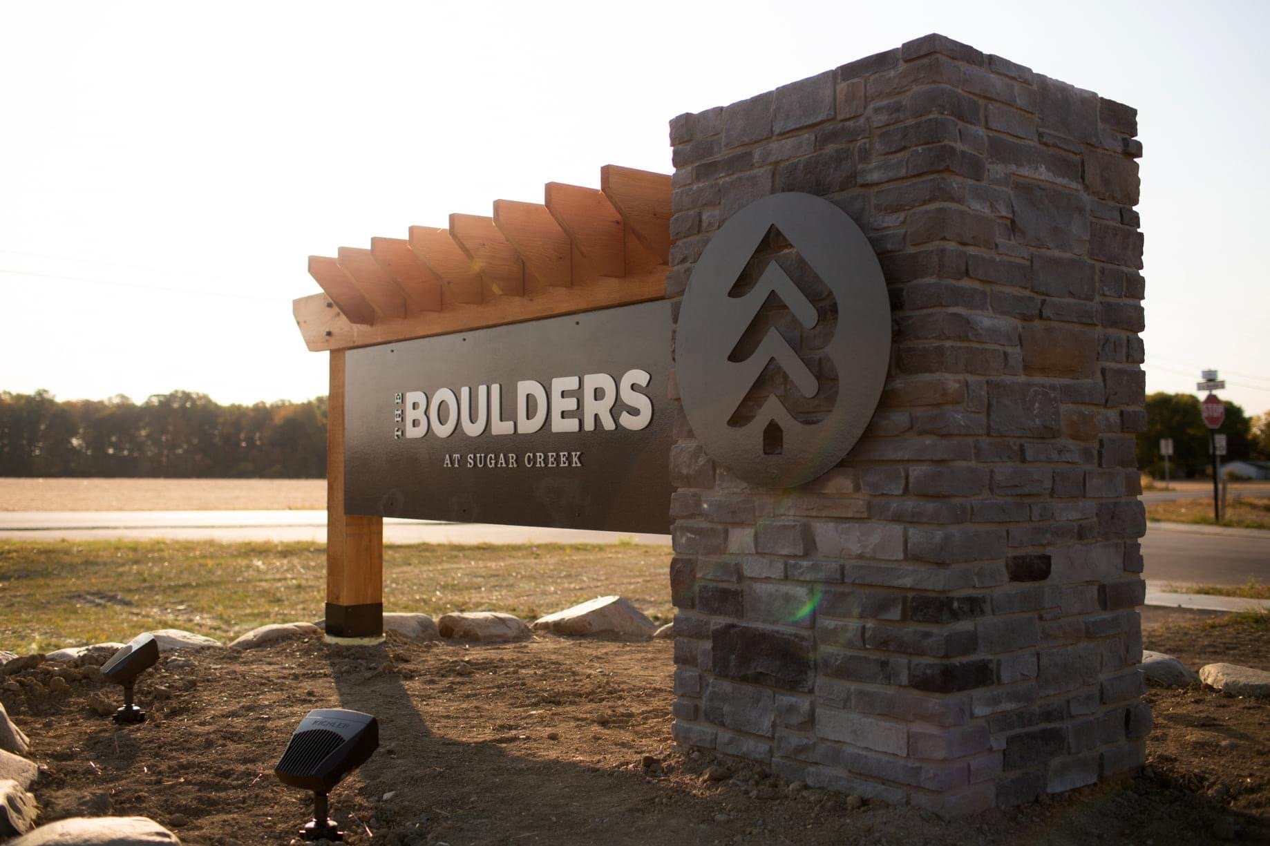 Sign at Boulders at Sugar Creek with a brick pillar and metal logo, landscape background, and outdoor lighting