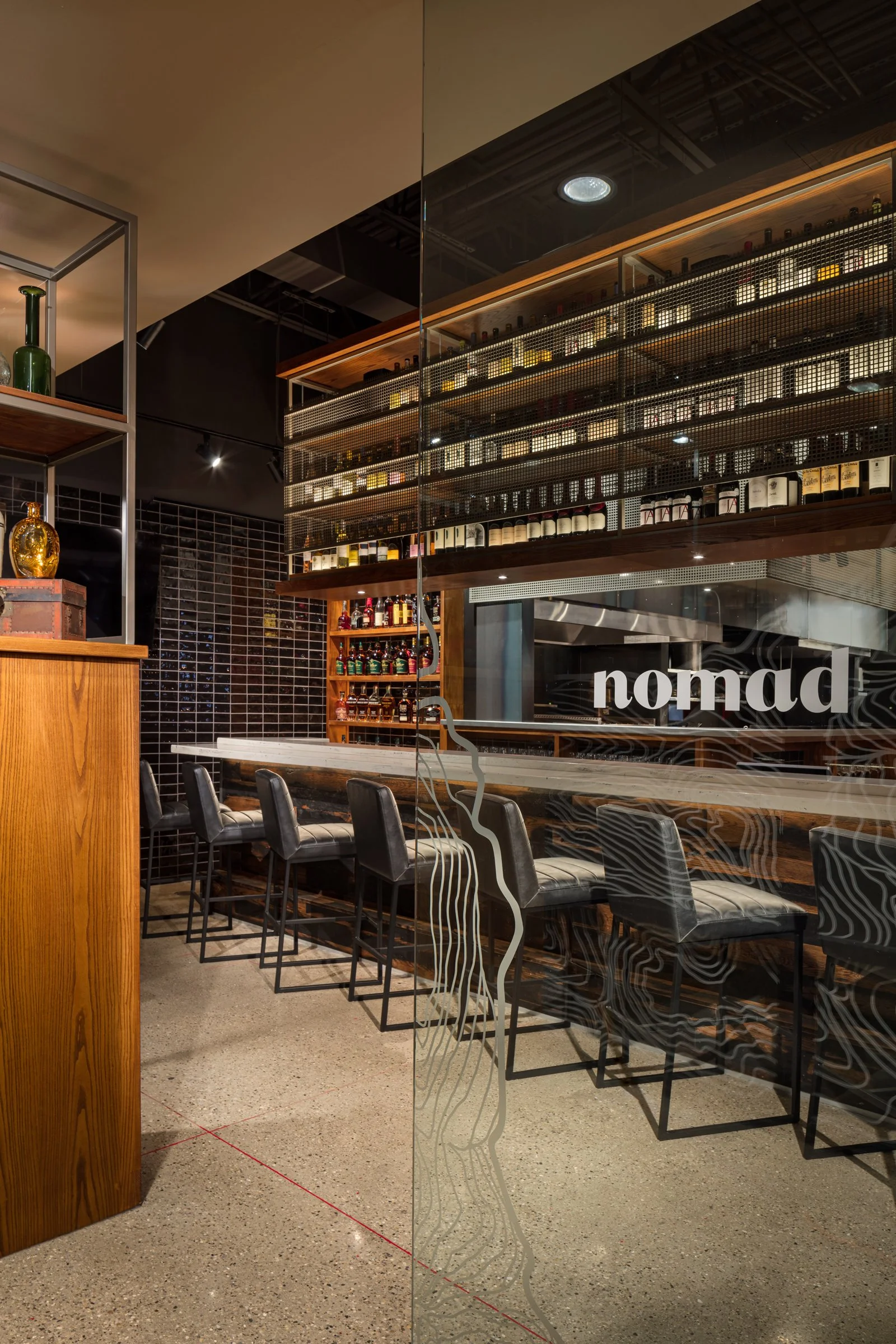 This image shows an interior of a modern bar or restaurant with a black tile wall, shelves filled with bottles, and a row of black leather bar stools along a counter. The reflection of the 
