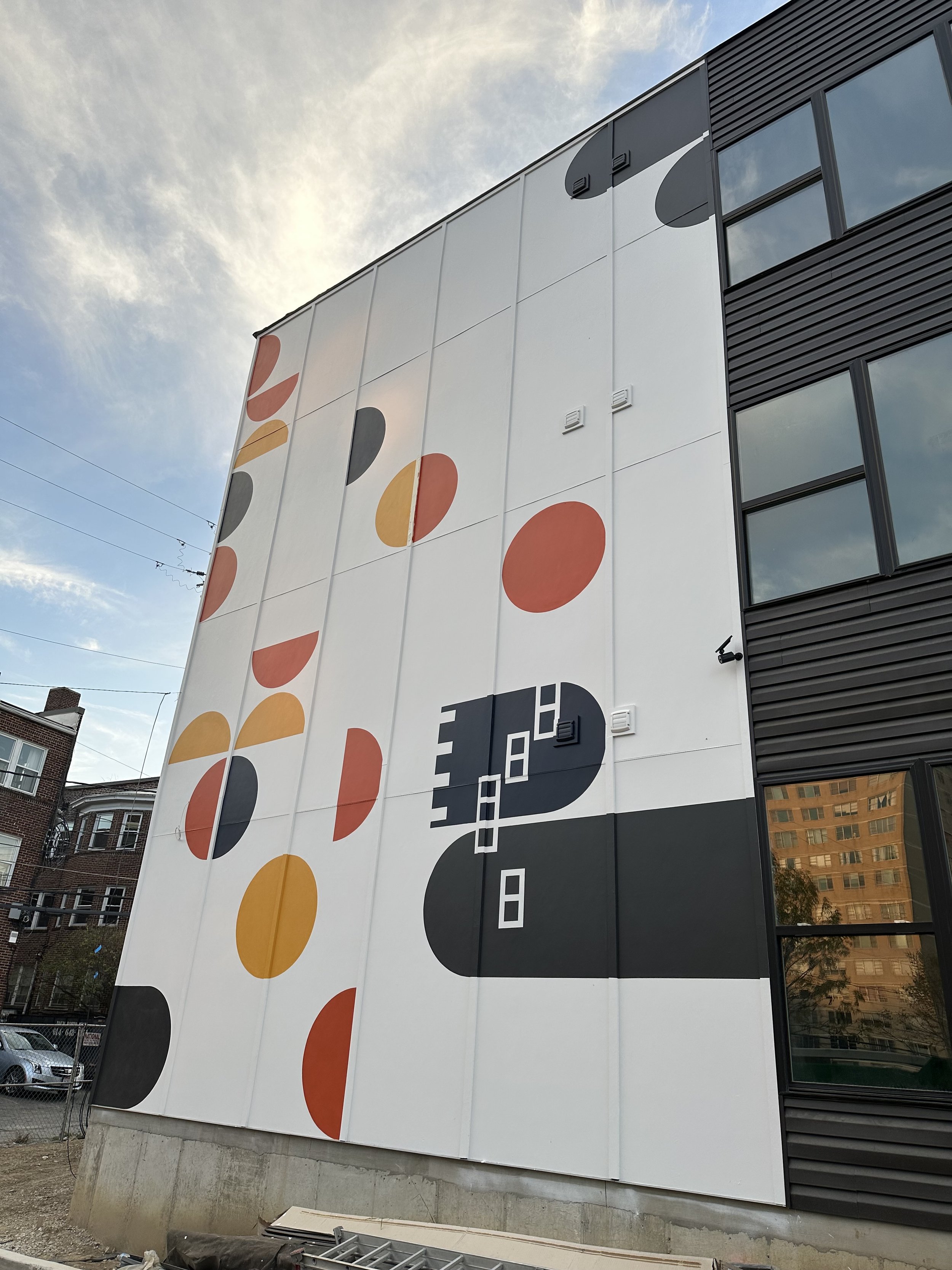 Large building with modern black and white exterior, featuring colorful abstract circular and geometric mural, with windows and reflections of trees and neighboring buildings.