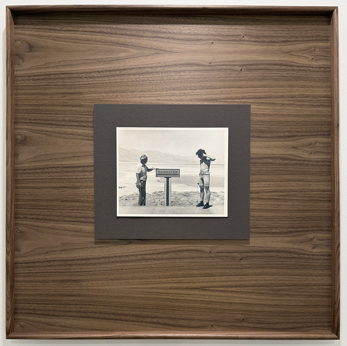 Black and white photograph of two people standing on a beach, one is holding a sign that says "BADWATER". The person on the left is wearing a hat and plaid shirt, the person on the right has short hair and is in casual clothes. Mountains and water ca