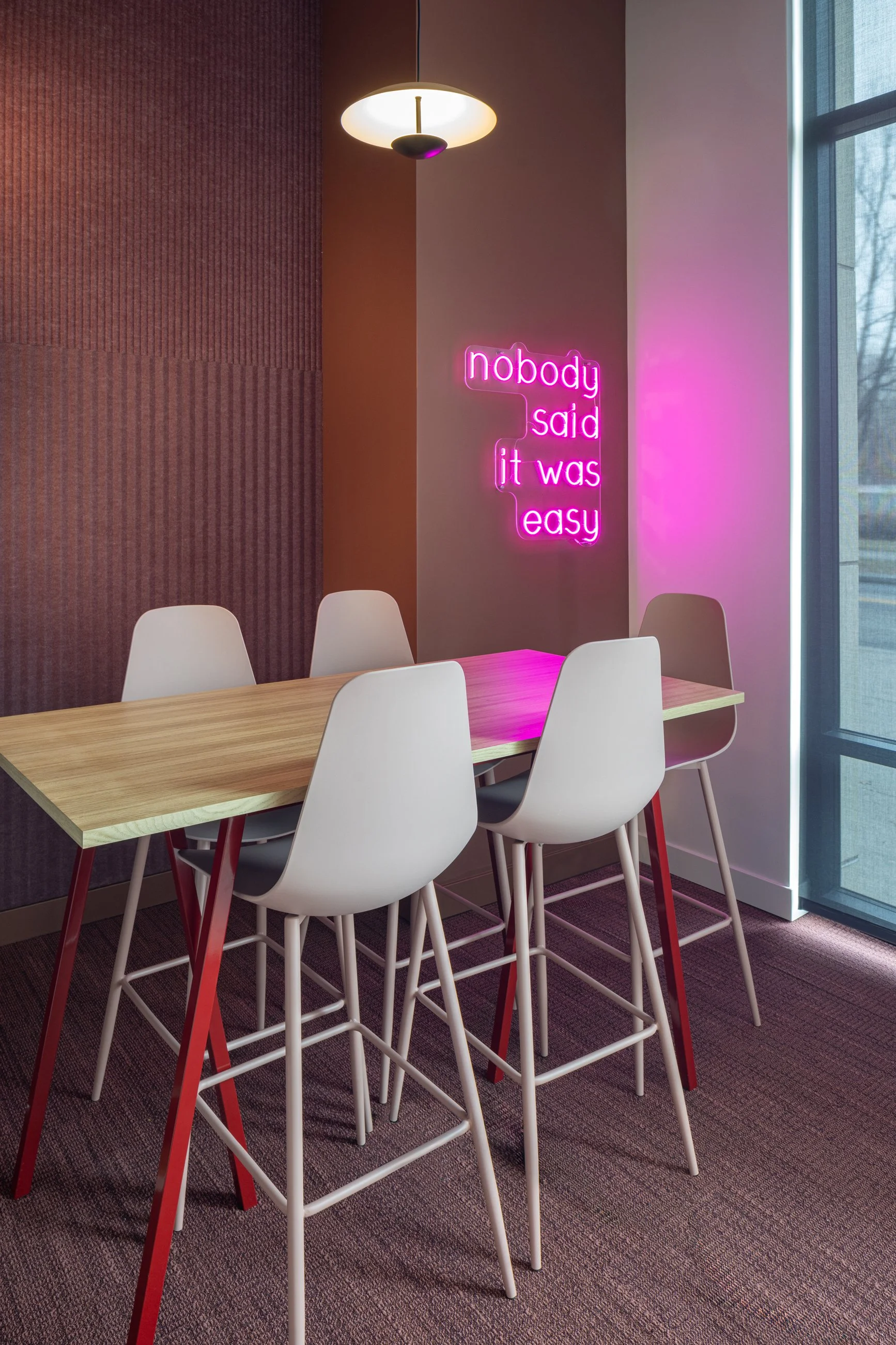 A modern dining area with a wooden table and five white chairs, a pink neon sign on the wall says 'nobody said it was easy', a pendant light hanging from the ceiling, and a large window with a view outside.