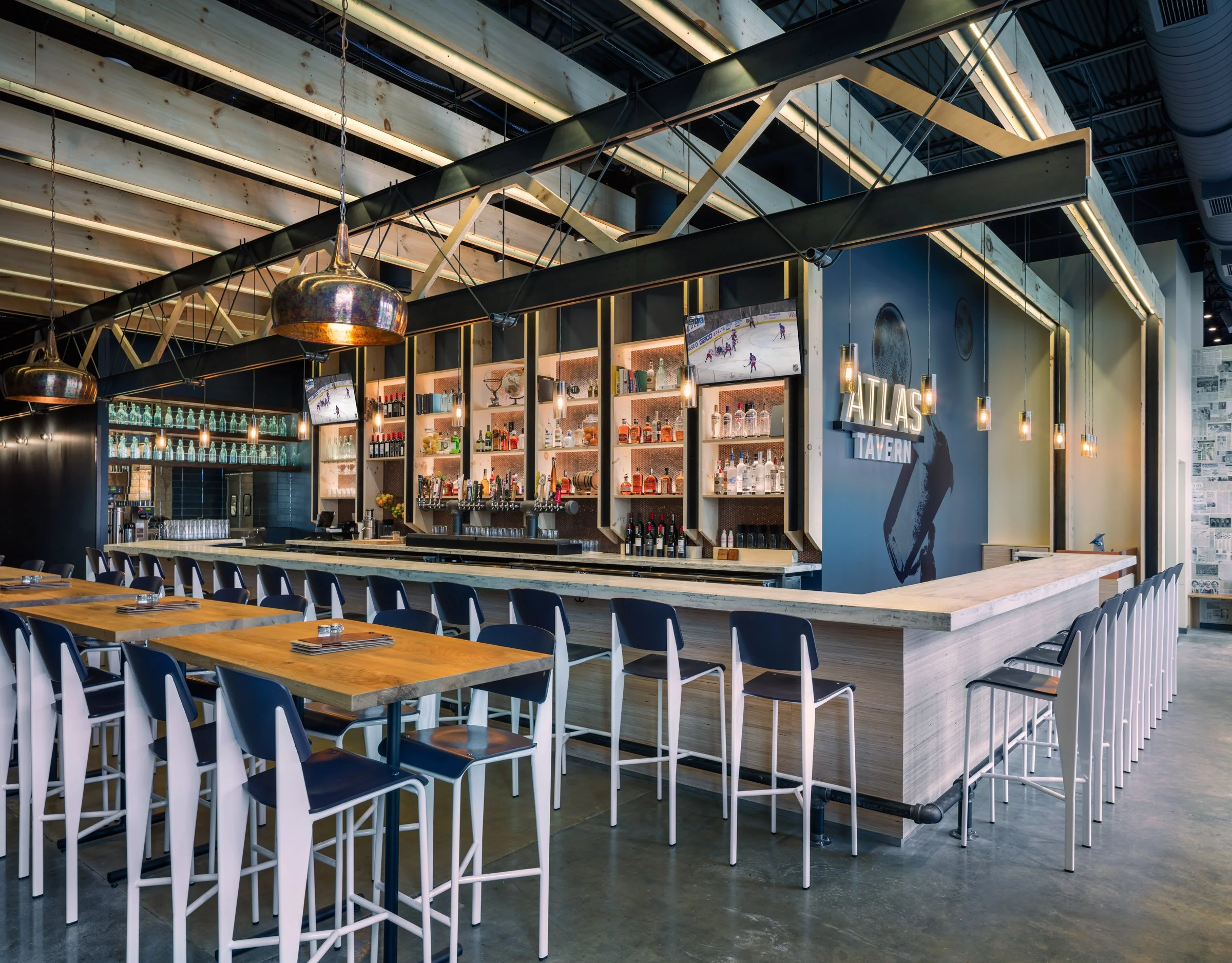 Interior of a modern bar with a long wooden counter, bar stools, and an overhead shelving unit stocked with bottles and glasses, with a sign that says 'Atlas Tavern' on the wall.