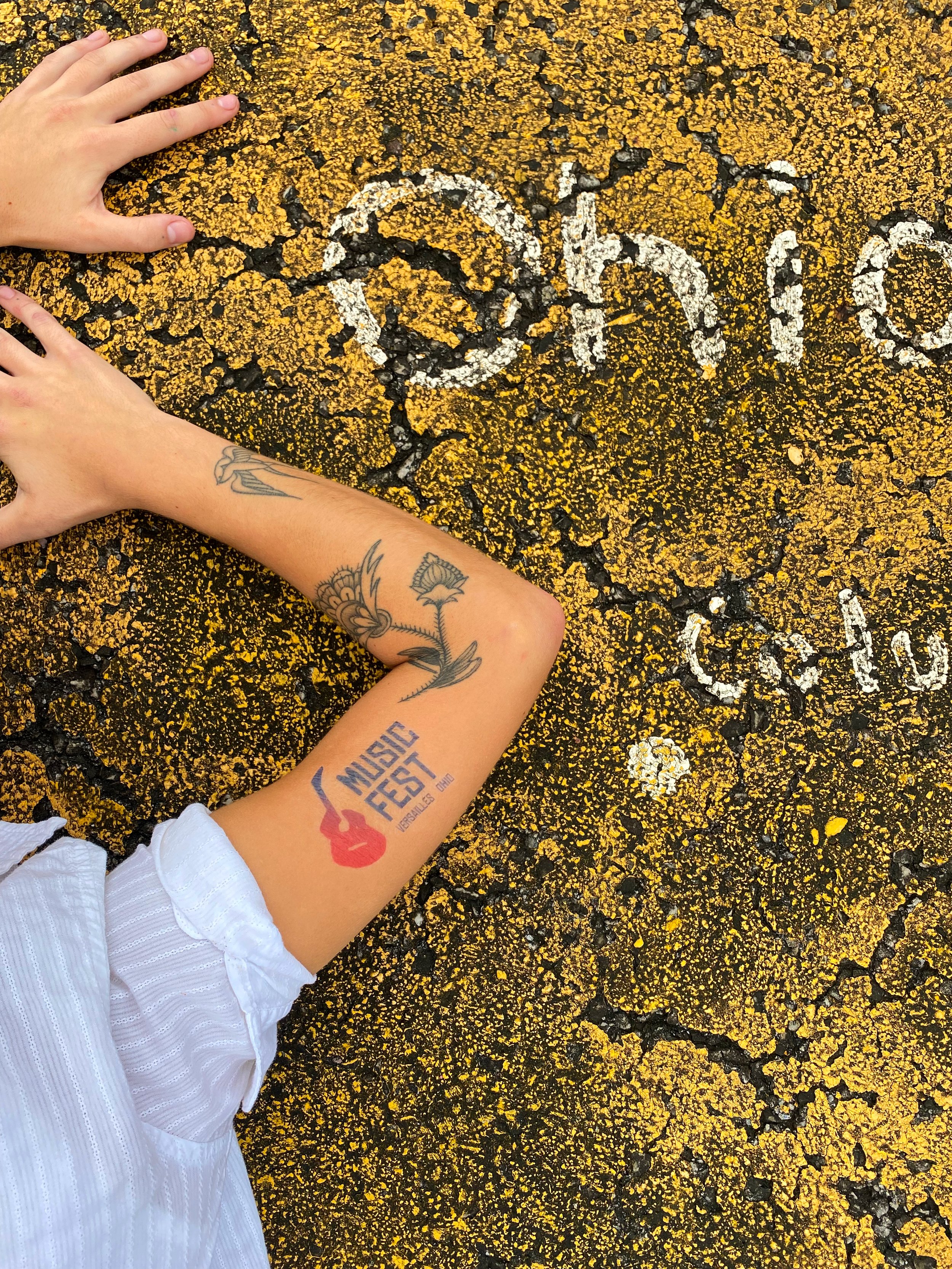Close-up of a person's arm with tattoos resting on cracked yellow and black pavement. The person is wearing a white sleeve. The pavement has white chalk writings that say "Music Fest" and partially visible "Oklahoma."