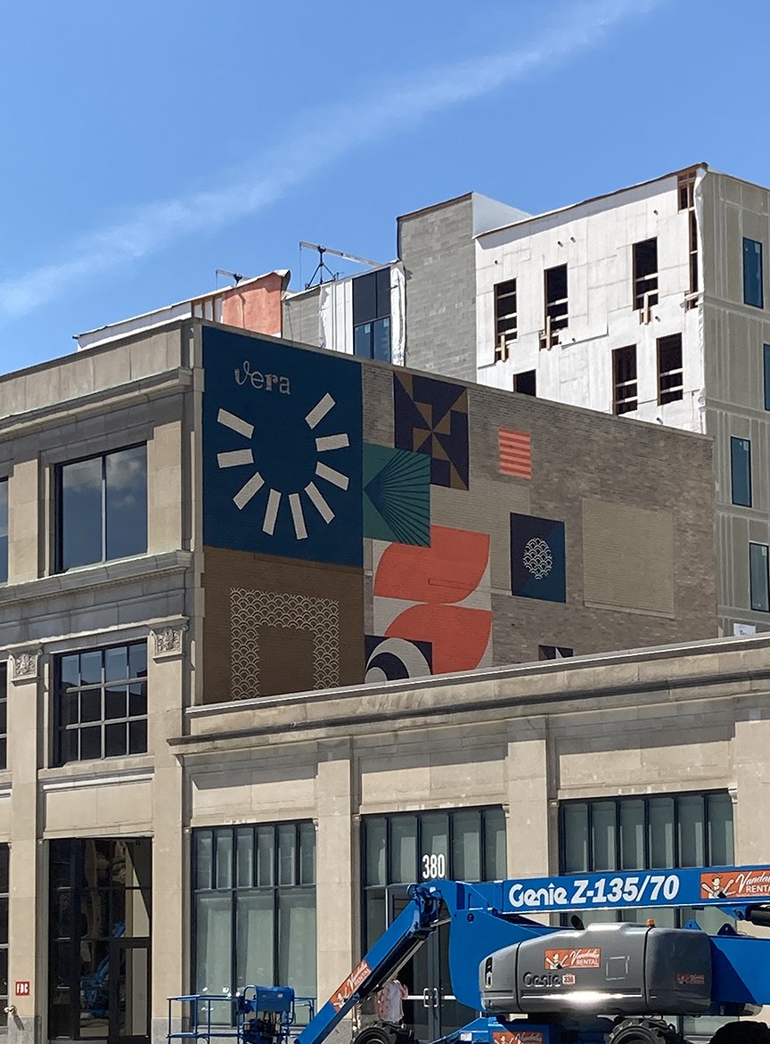 A multi-story building with a colorful mural on its side, featuring geometric shapes and patterns, with a clear blue sky above and construction equipment in the foreground.