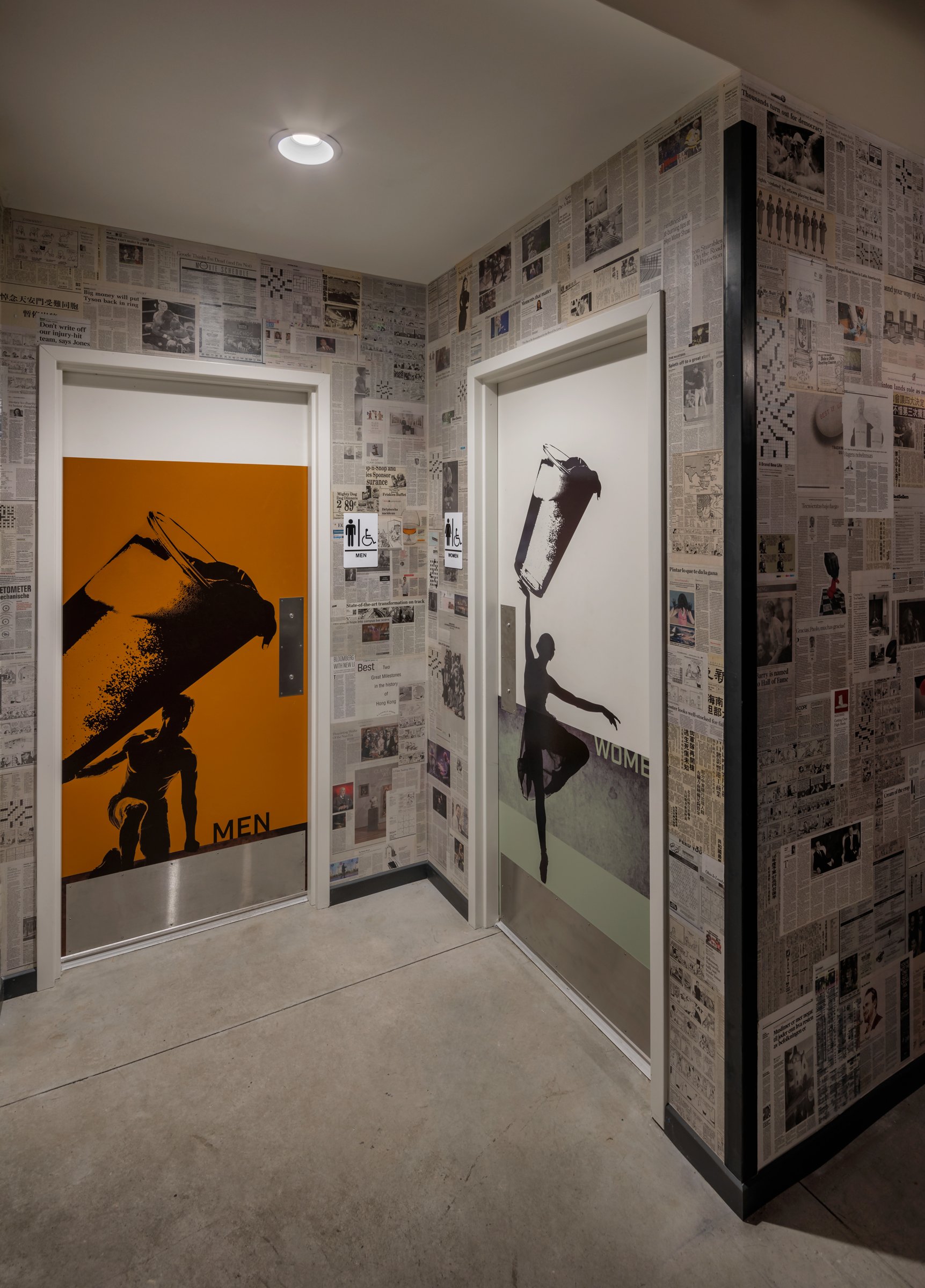 Two elevator doors with artistic images; the men's door has an orange background with a black silhouette of a person lifting a large object, with the word 'MEN'; the women's door features a white background with a silhouette of a ballerina holding a 