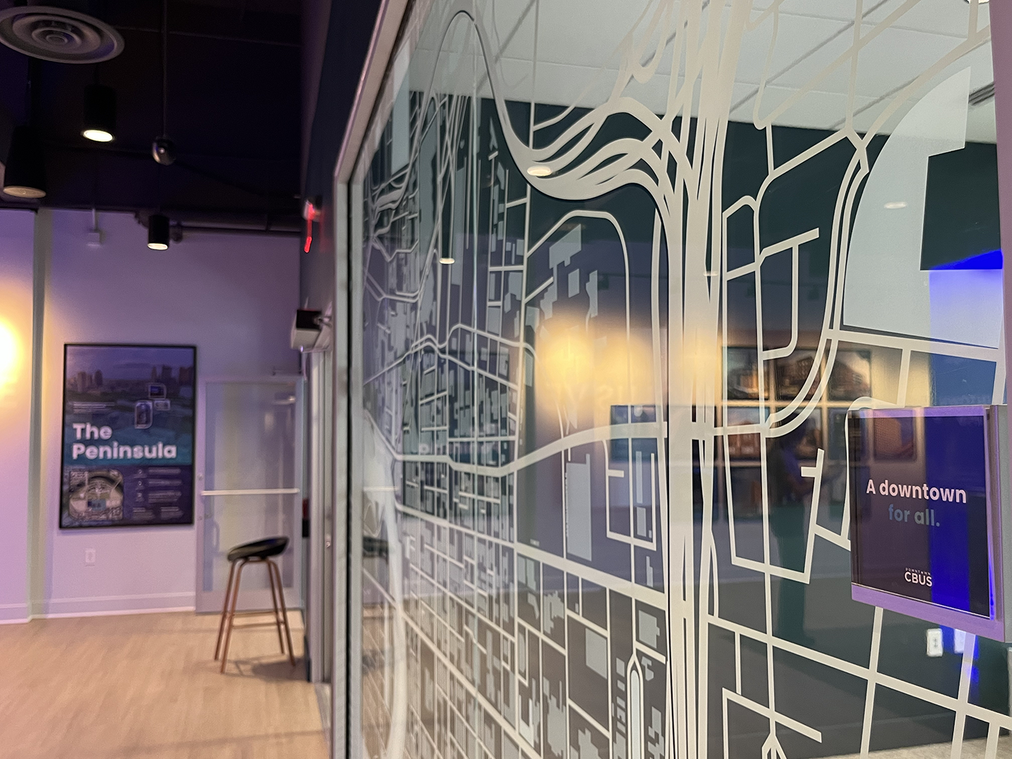 Interior of a modern office with a large glass wall featuring a city map design, a promotional poster on the wall that reads "The Peninsula," and a TV screen that displays "A downtown for all."