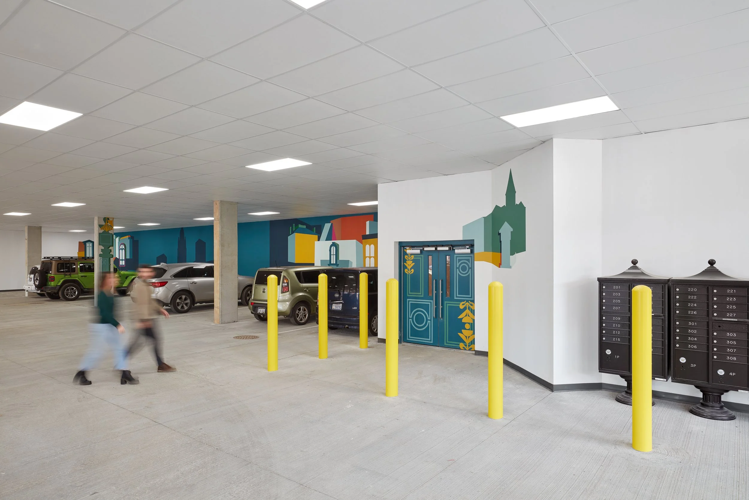 Underground parking garage with parked cars, colorful wall mural, yellow security posts, and black mailboxes.