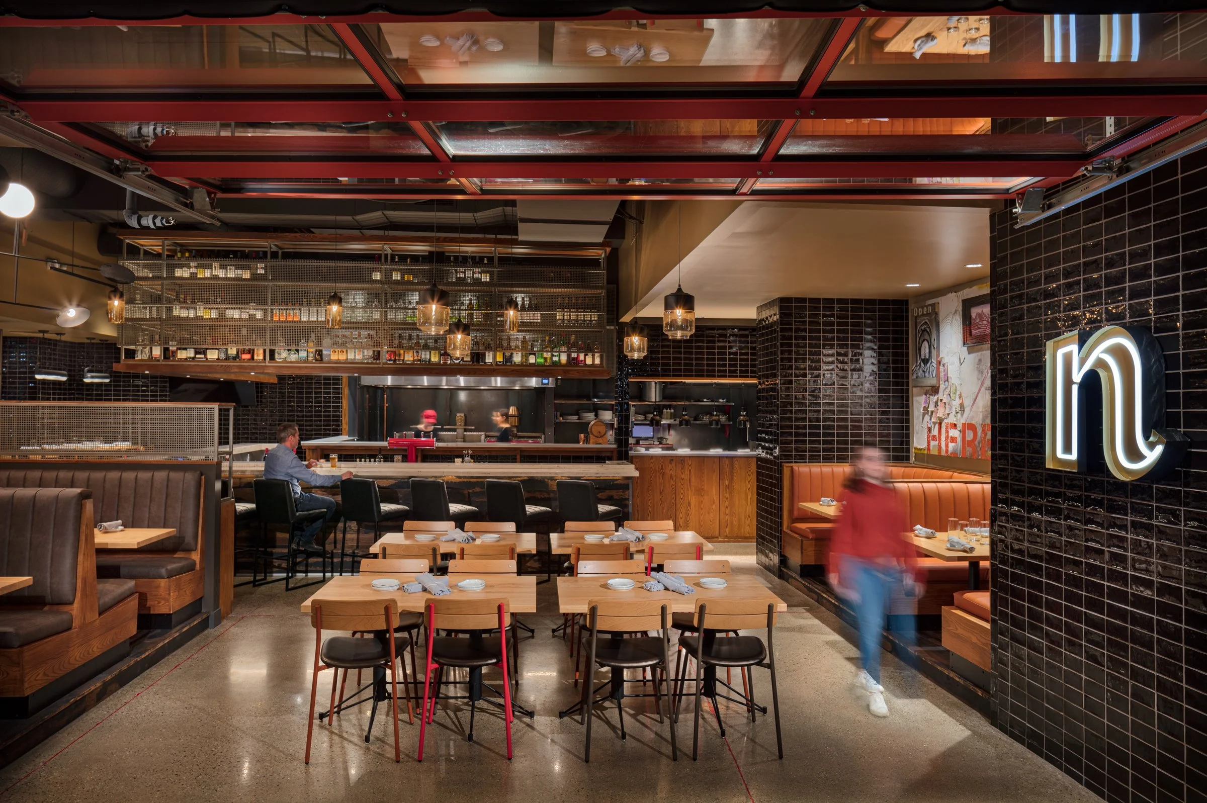 Interior of a modern restaurant with wooden tables, black and brown chairs, black tiled walls, and a visible open kitchen in the background. Pendant lights hang from the ceiling, and a neon sign displays a stylized 'n' on the wall.