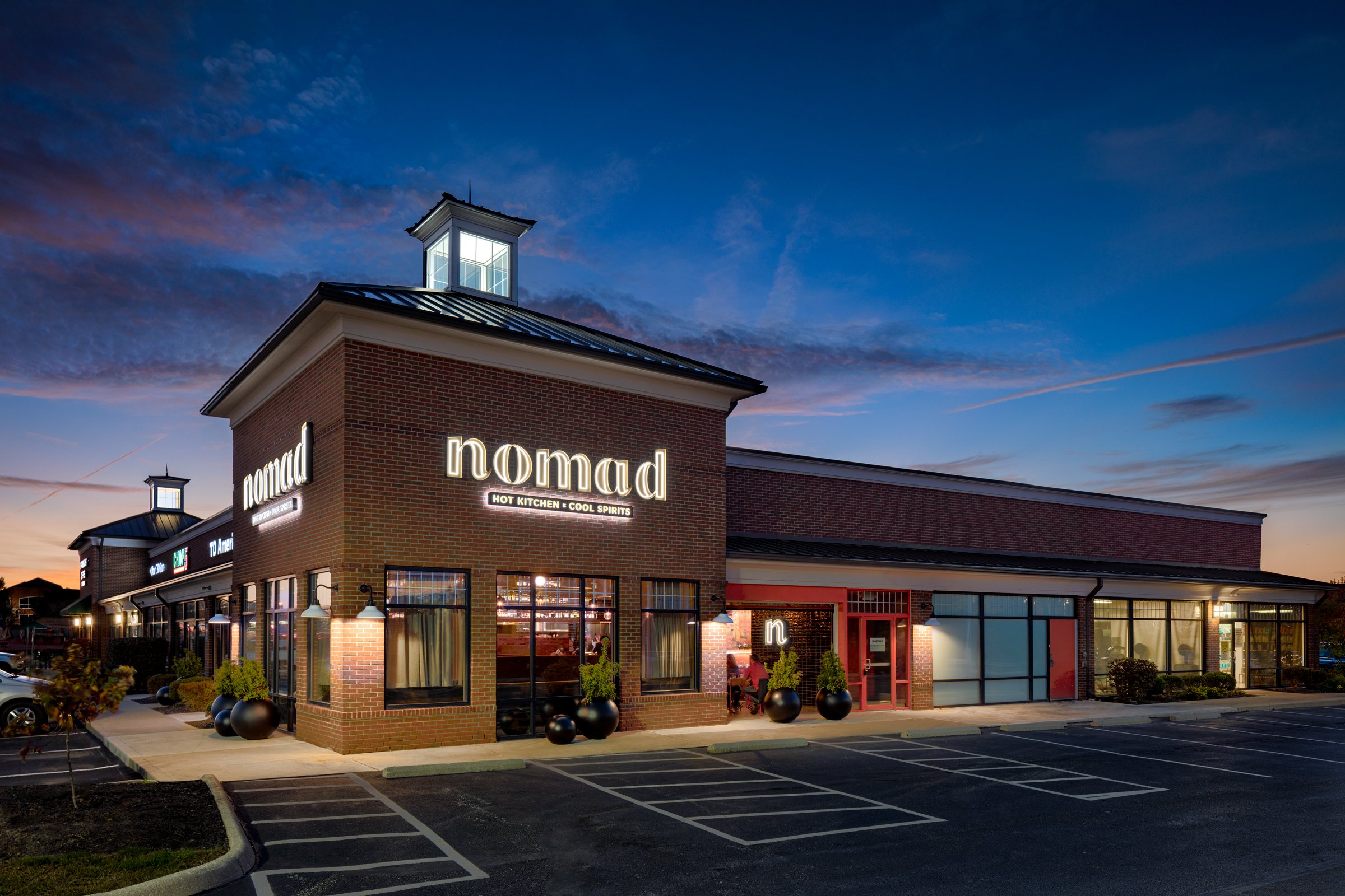 Exterior of a brick restaurant called 'Nomad' during dusk, with illuminated signs, large windows, and a parking lot in front.