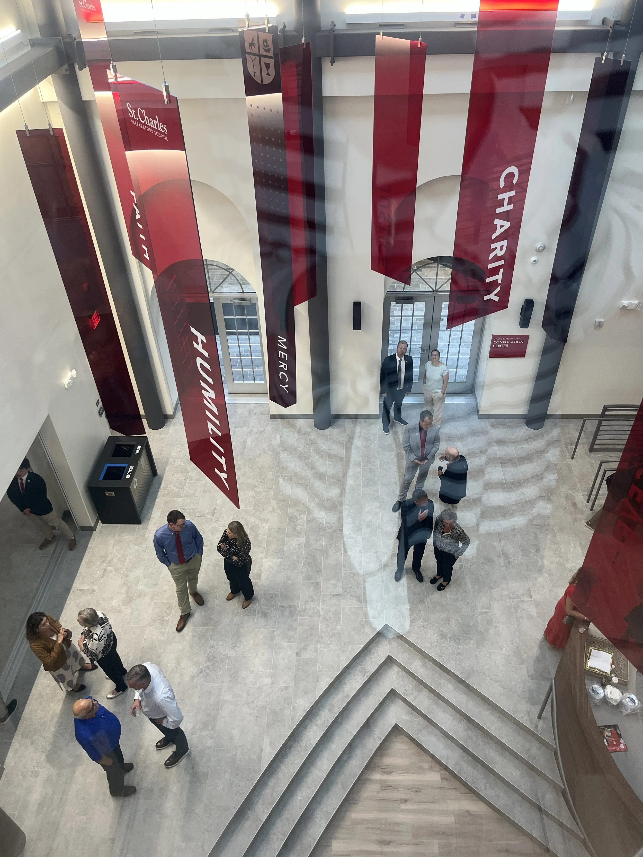 People gathered in a spacious, well-lit lobby with large red banners hanging from the ceiling that have British flags and the words 'Charity,' 'Humility,' and 'Mercy.' The view is from above, showing an entrance with glass doors and windows.