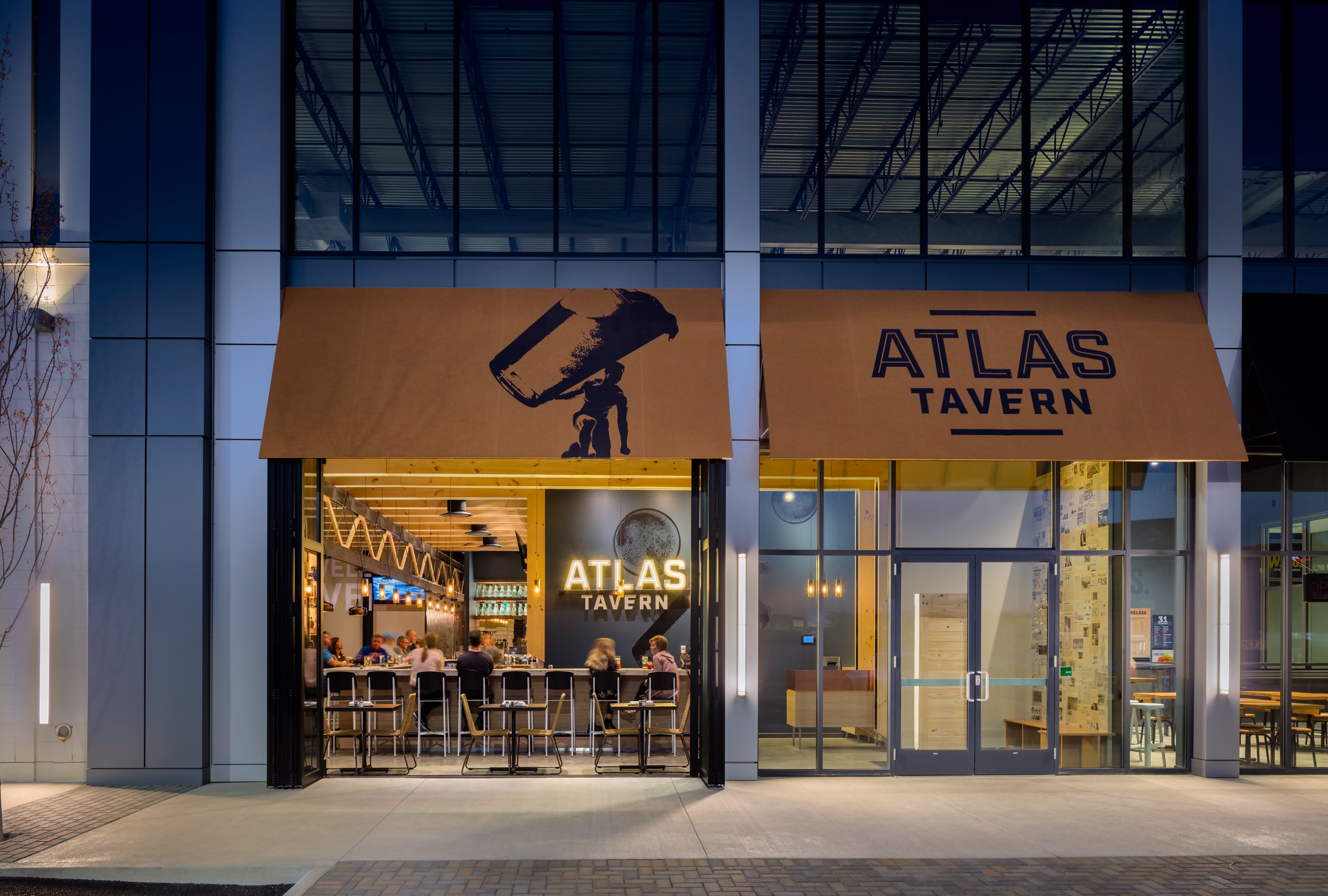 Exterior view of Atlas Tavern restaurant with large signage, open front bar area with patrons, modern design, evening lighting.