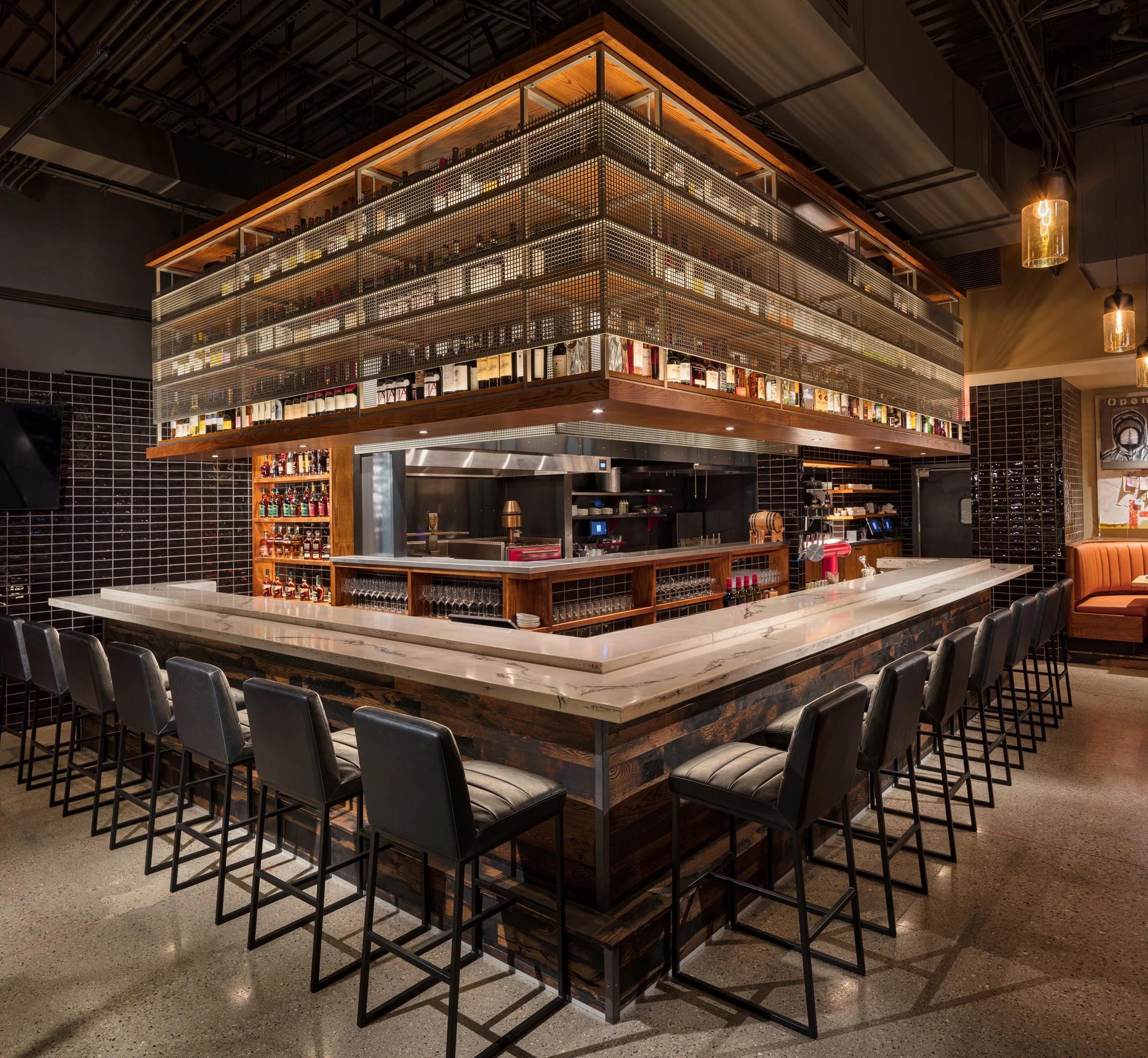 Modern bar with marble countertop and black leather stools, surrounded by liquor bottles and glassware, with a suspended liquor shelf above.